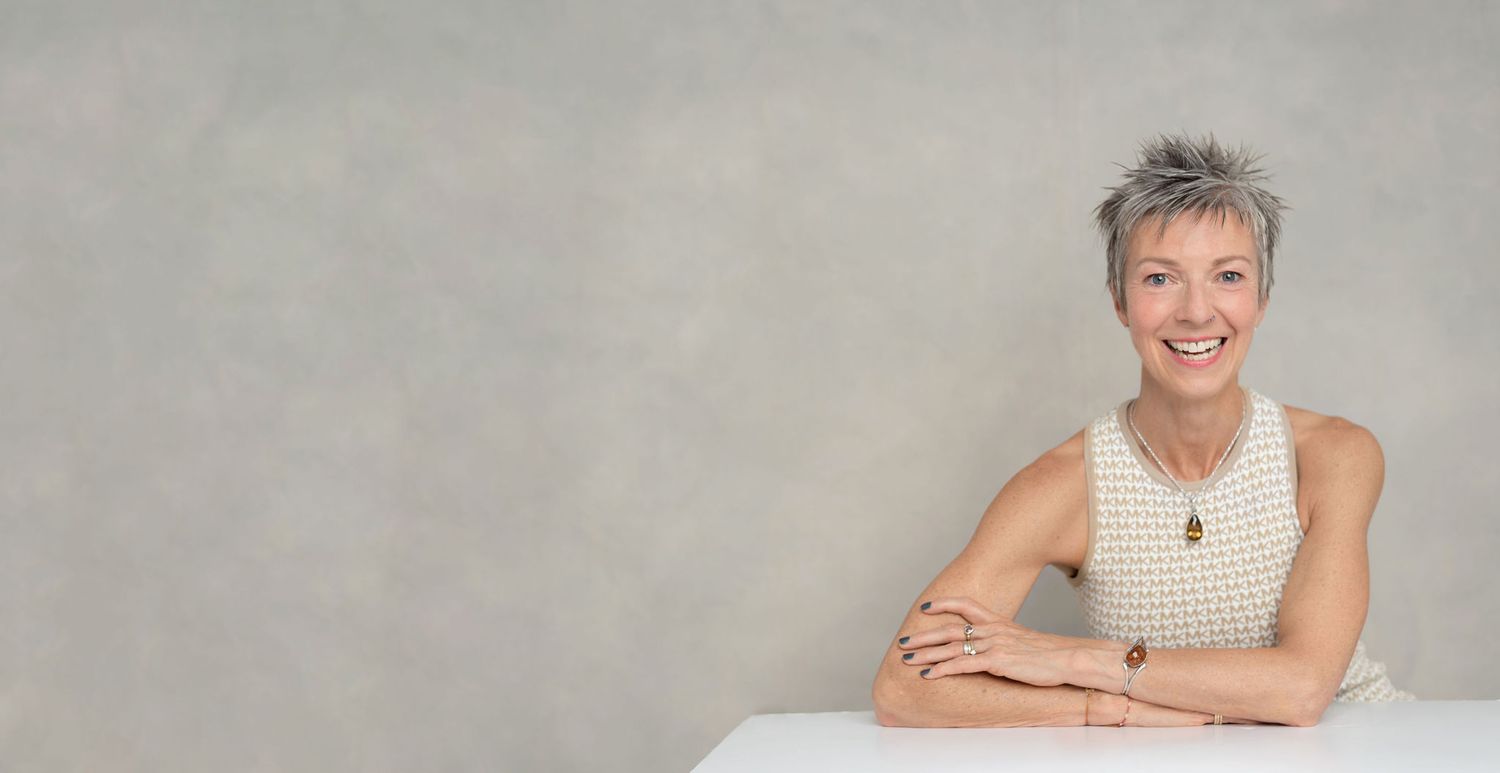 Casual professional portrait showing relaxed pose in white sleeveless top against grey background.