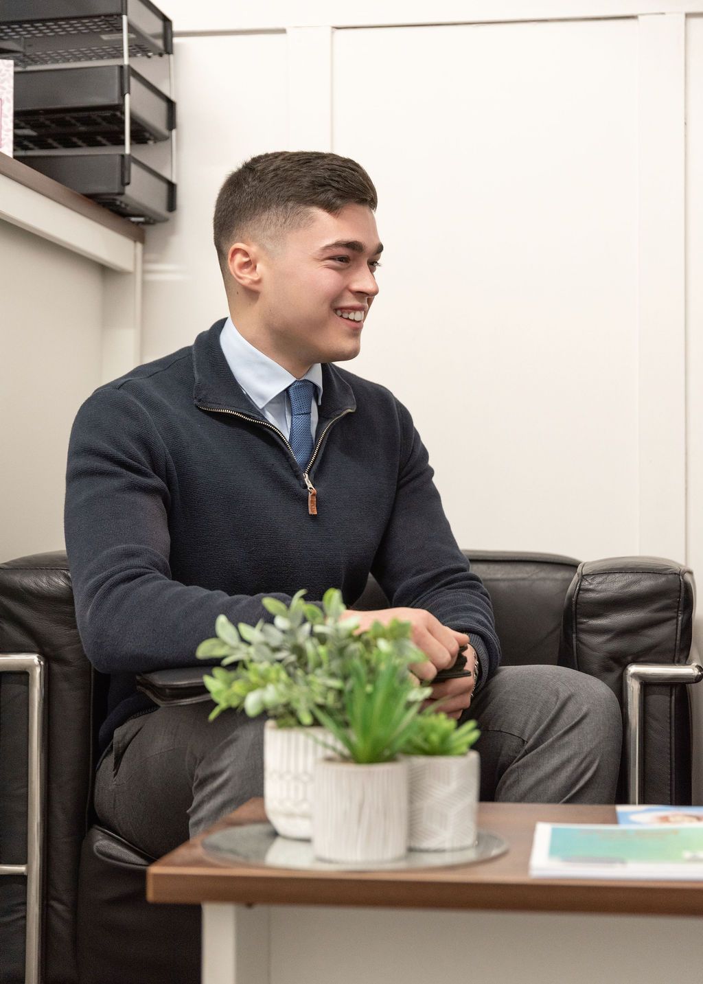 Professional in a dark suit sits at a desk with a small potted plant in a modern office setting.