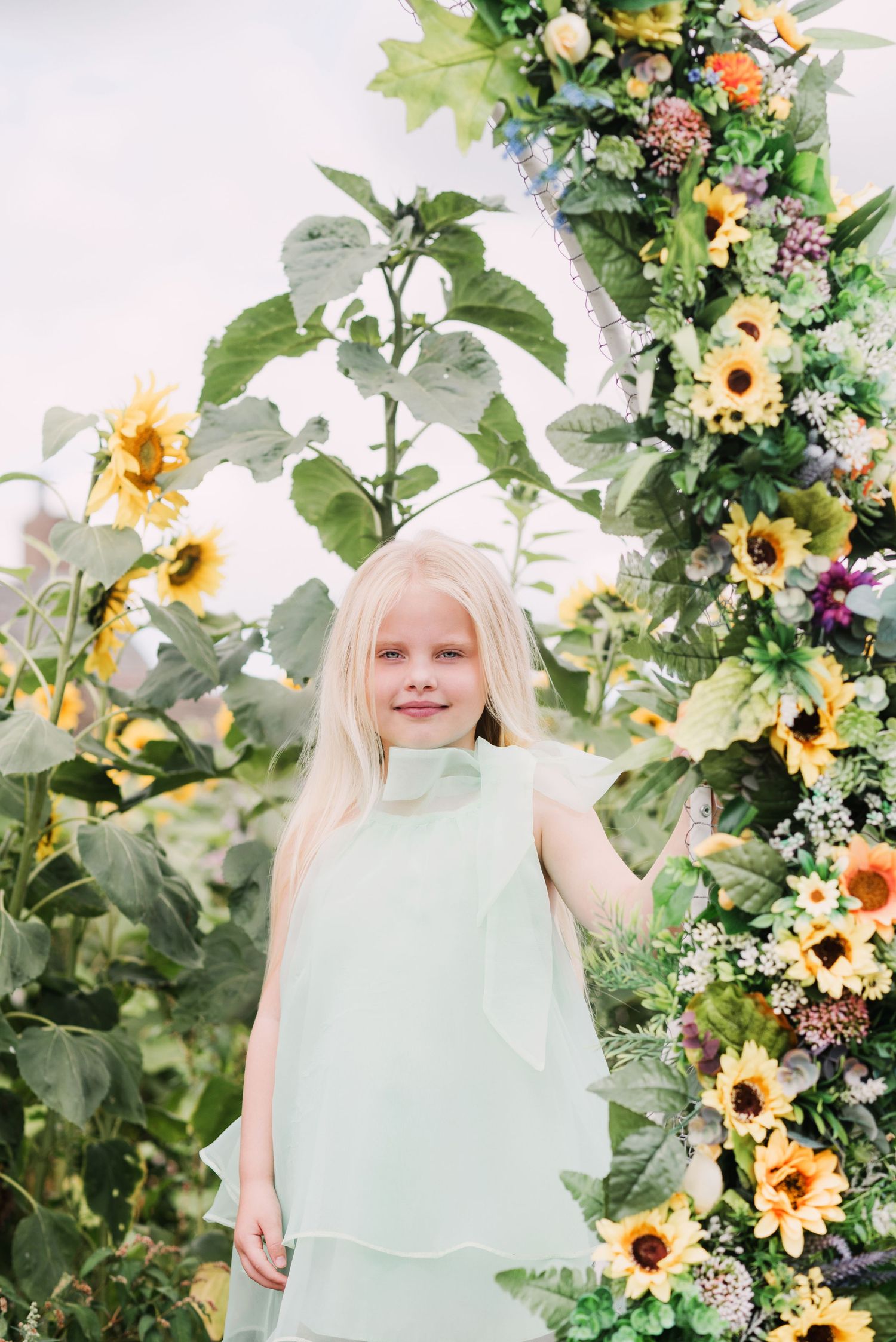 Young blonde girl in mint green dress smiling among sunflowers and floral arch outdoors.