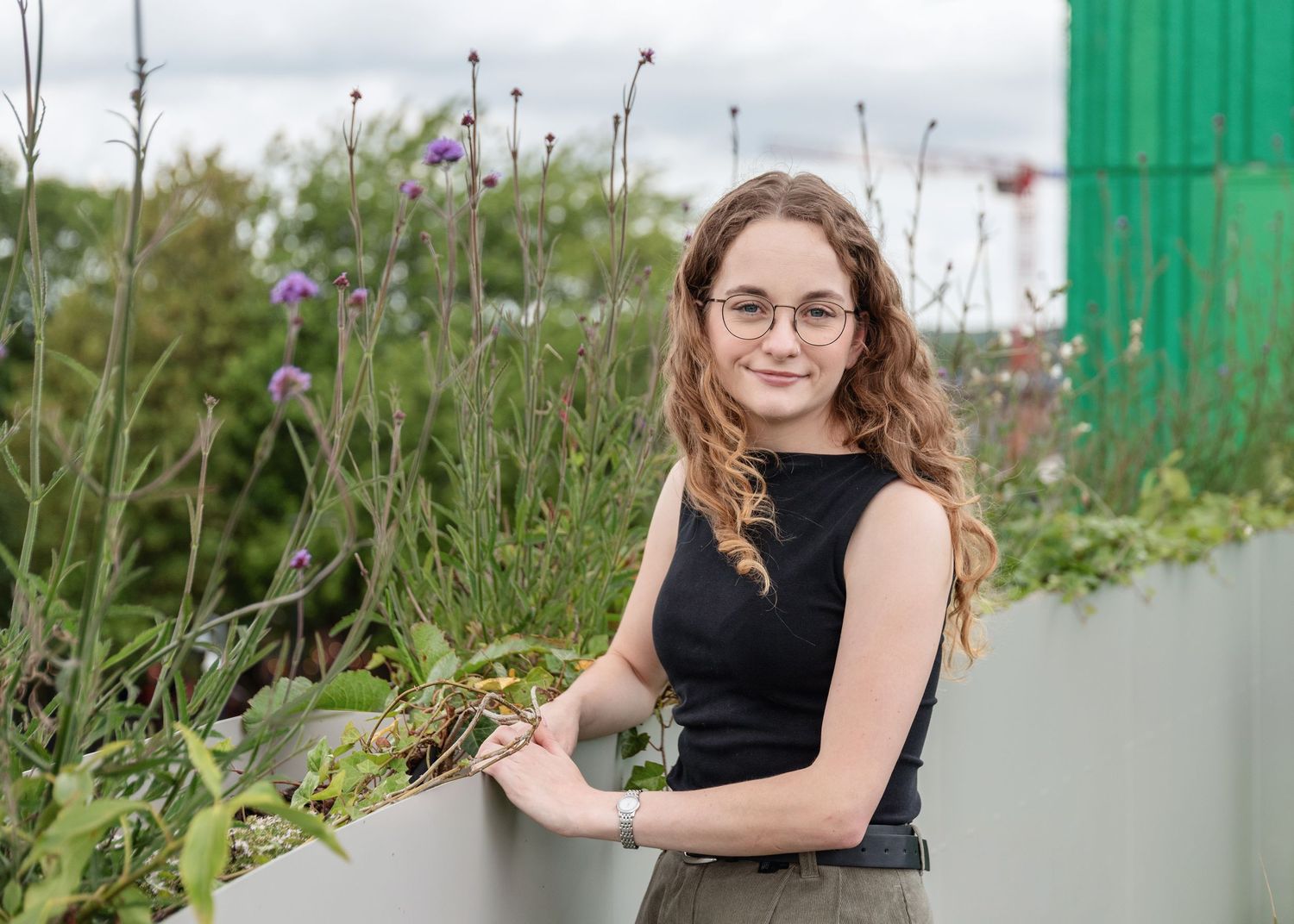 Young woman with curly hair and glasses smiling beside a rooftop garden with wildflowers and greenery.