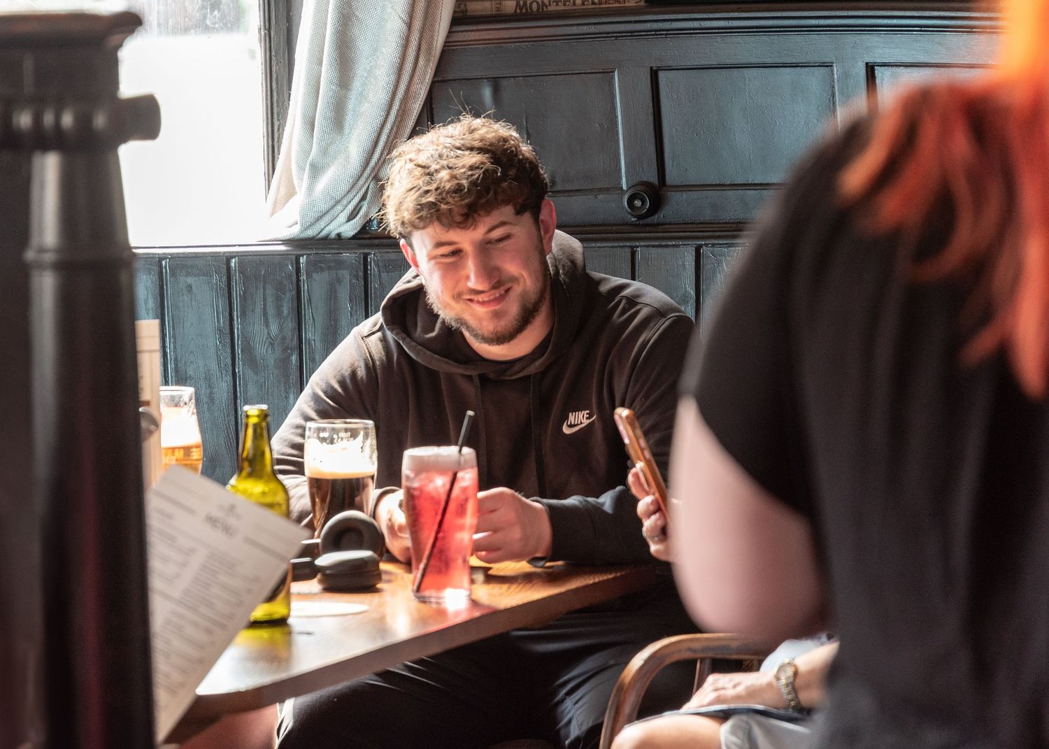 Young man in black Nike hoodie smiling at pub table with beer glasses, enjoying drinks with friends in cozy bar setting.