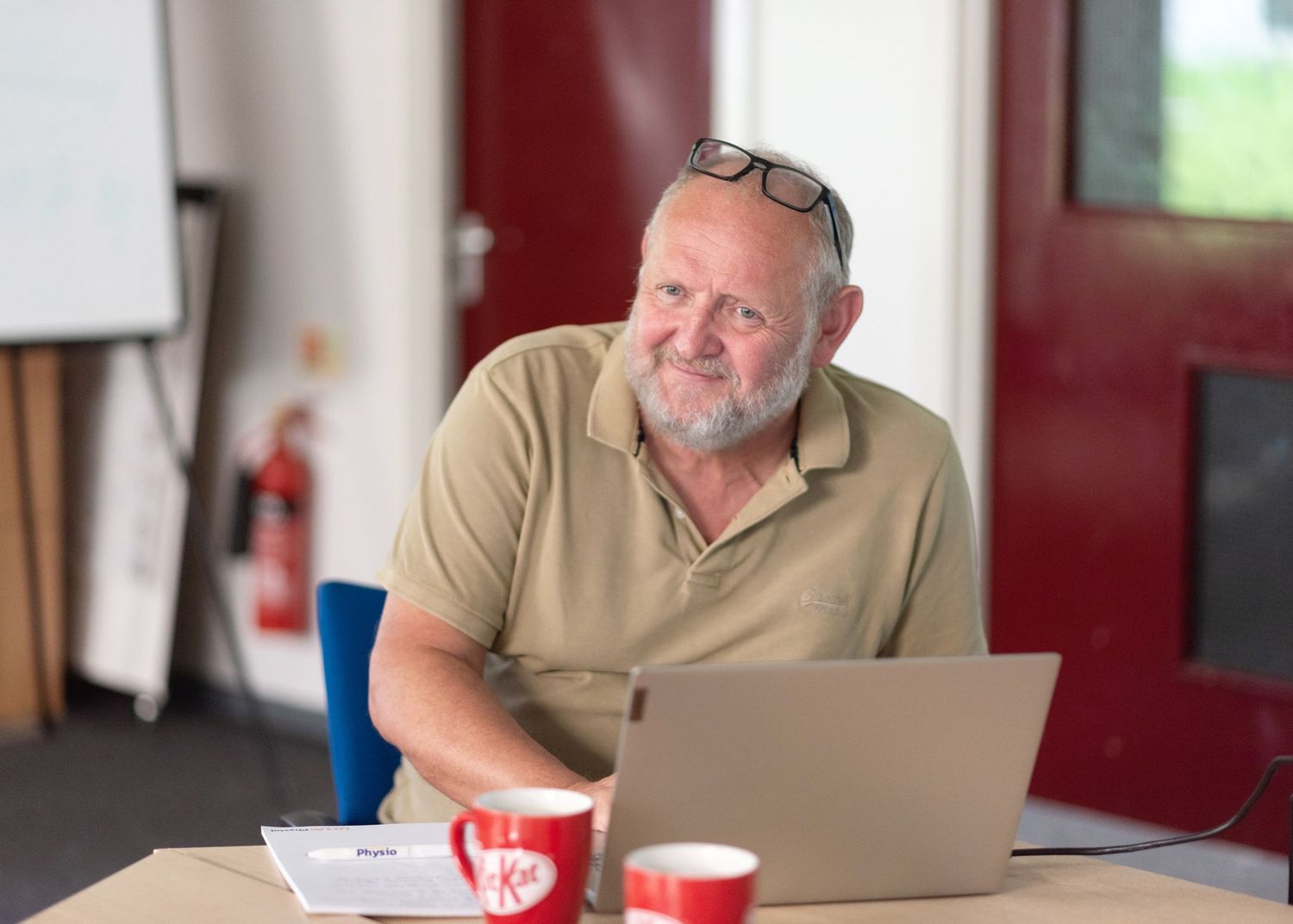 Smiling person wearing tan shirt works on laptop computer at desk with red cup in casual office setting.