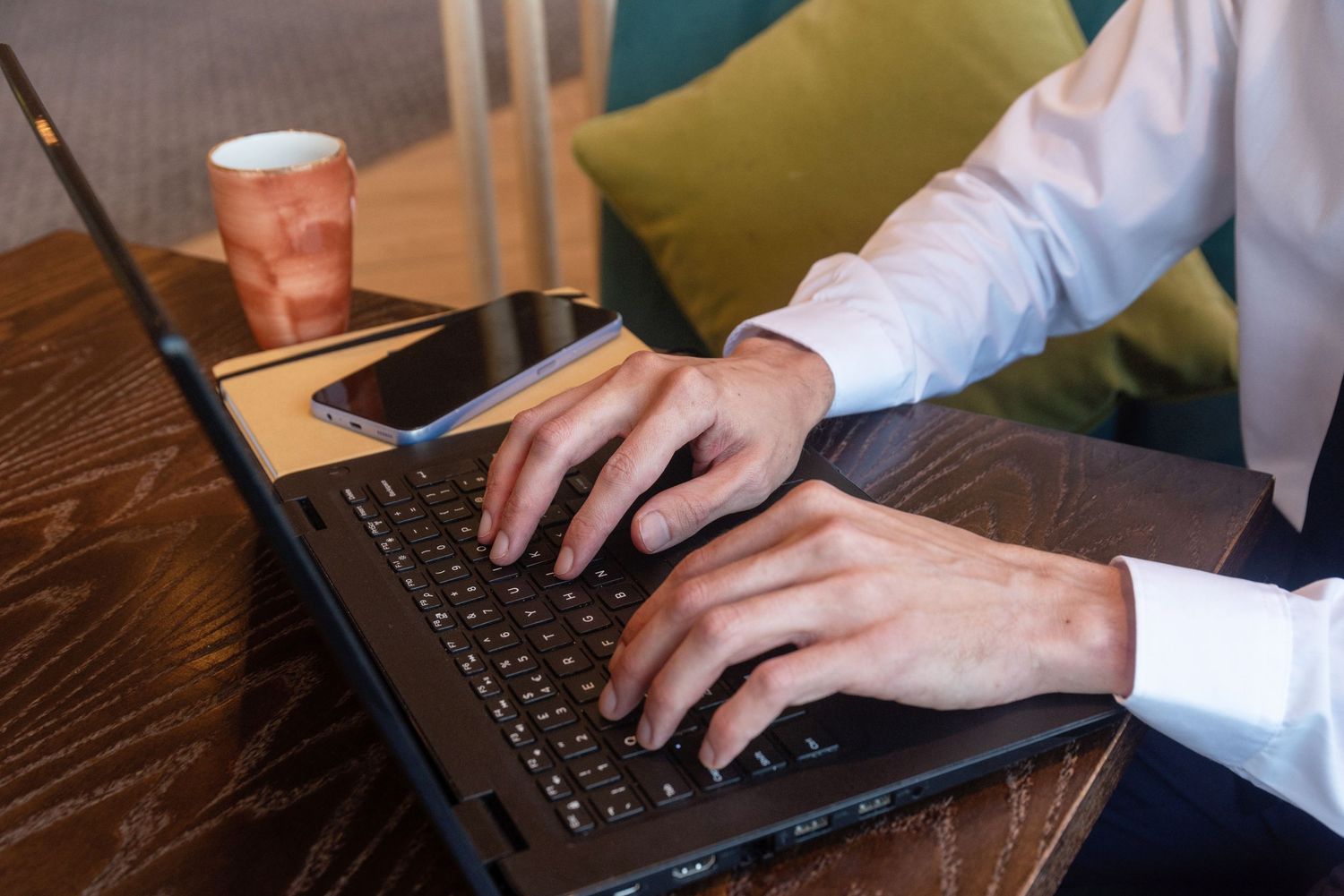 Hands typing on a laptop keyboard with a disposable coffee cup on wooden table.