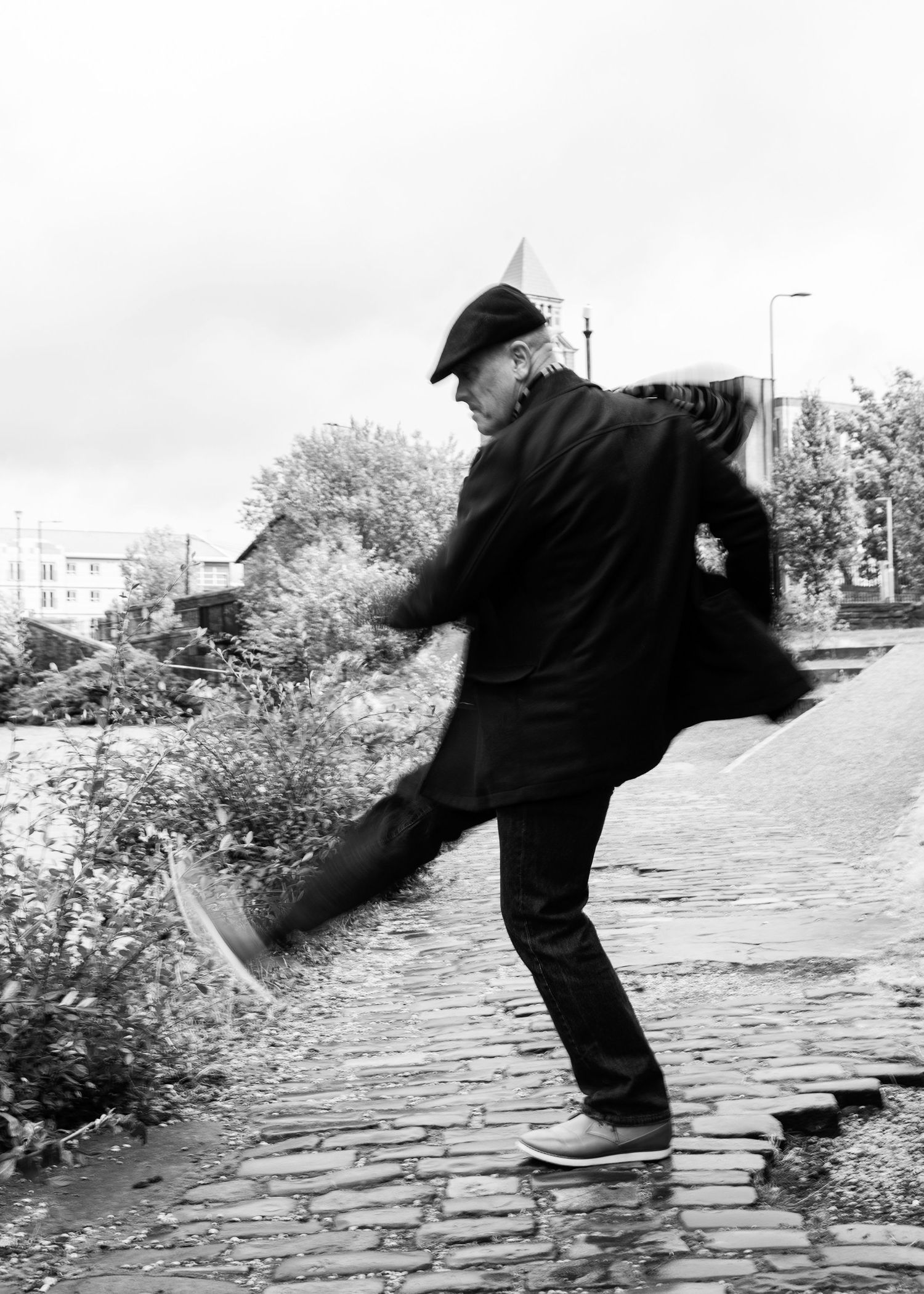A person in black clothing performs a playful kicking motion on a cobblestone path in a black and white photograph.