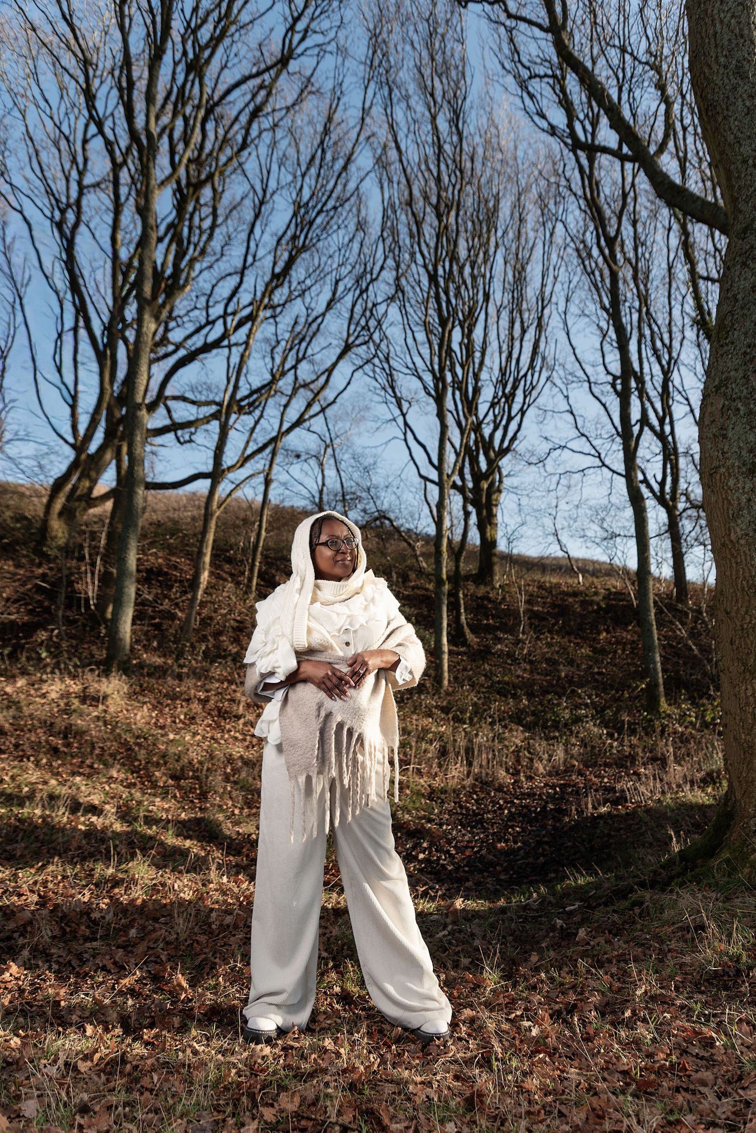 Person wearing a flowing white outfit dances gracefully among bare winter trees in a wooded hillside setting.