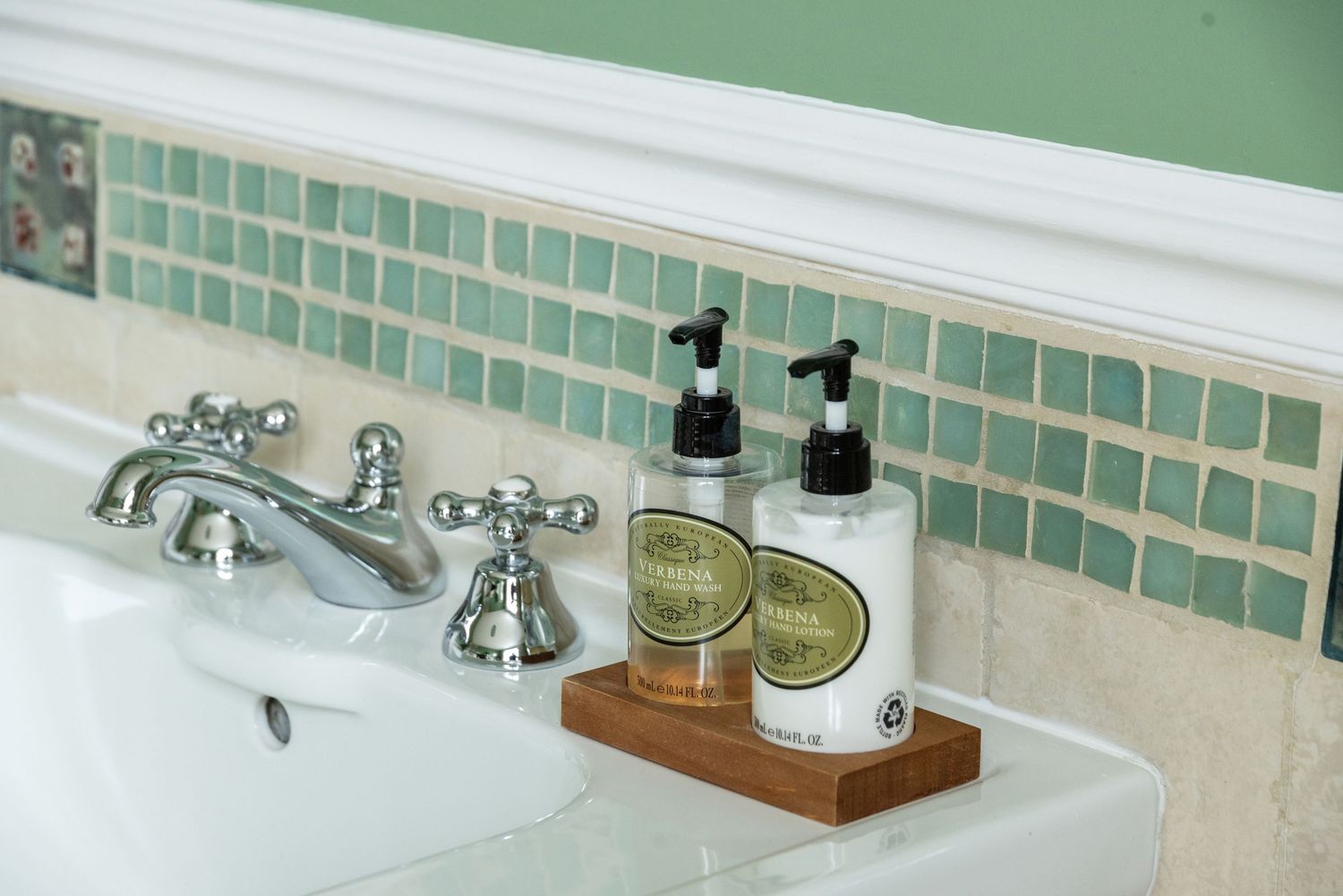 Elegant bathroom sink with chrome faucet and two pump soap dispensers on a wooden tray against green mosaic tiles.
