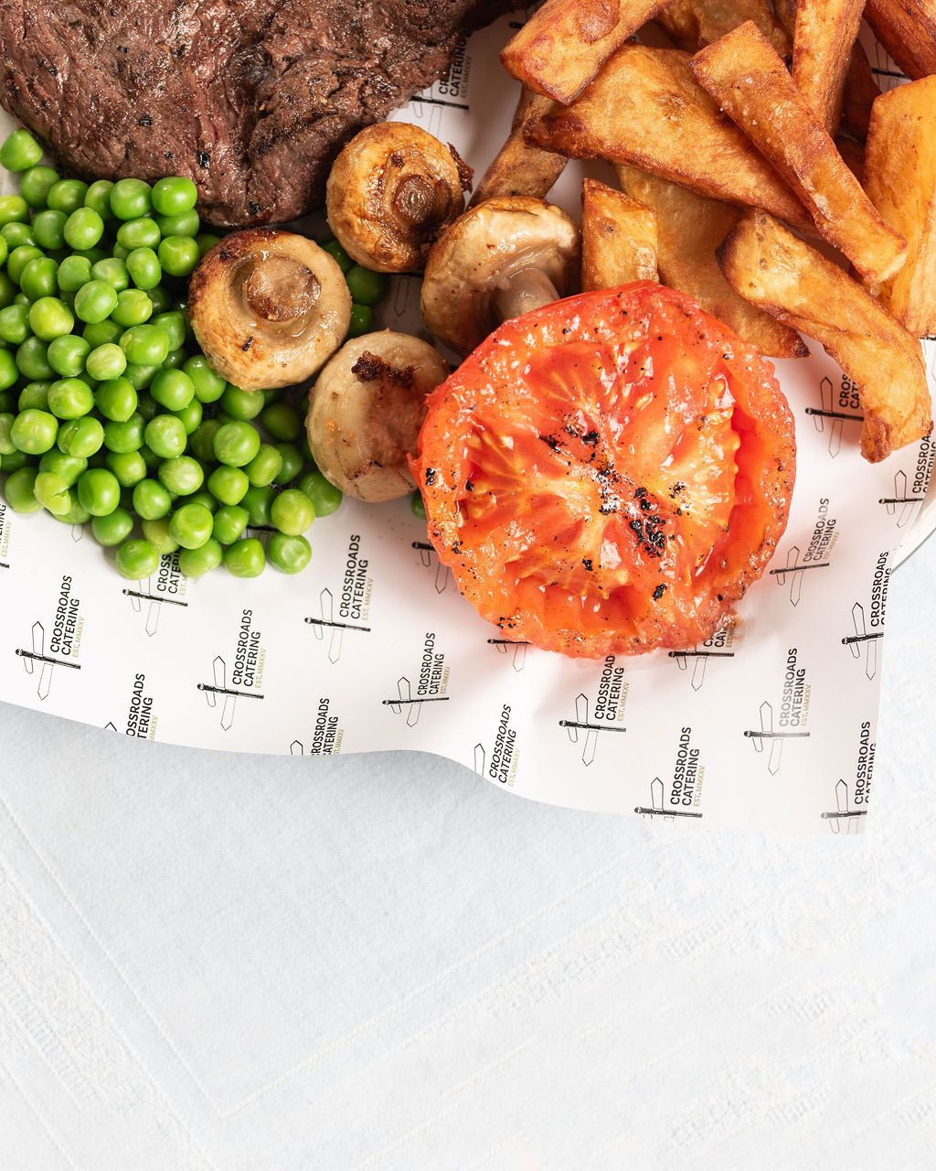 A hearty meal with grilled steak, peas, mushrooms, roasted tomato, and crispy potato wedges on branded paper.