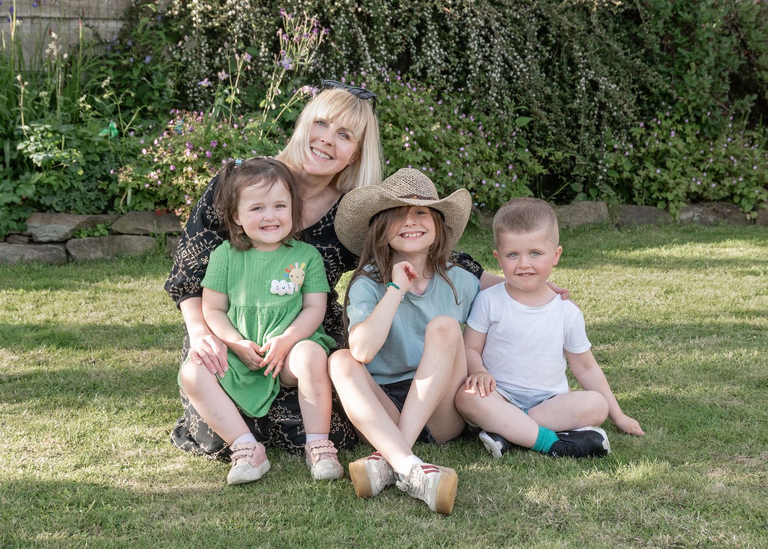 Woman with blonde hair sits on grass with three young children in a sunny garden setting.