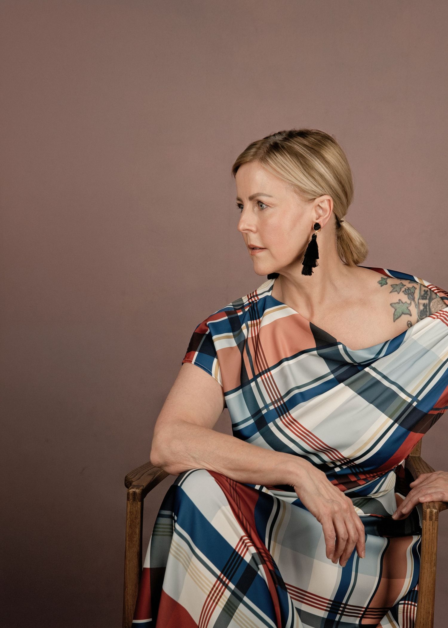 Woman in plaid dress with tattoo sits in wooden chair, wearing black earrings, against mauve background.