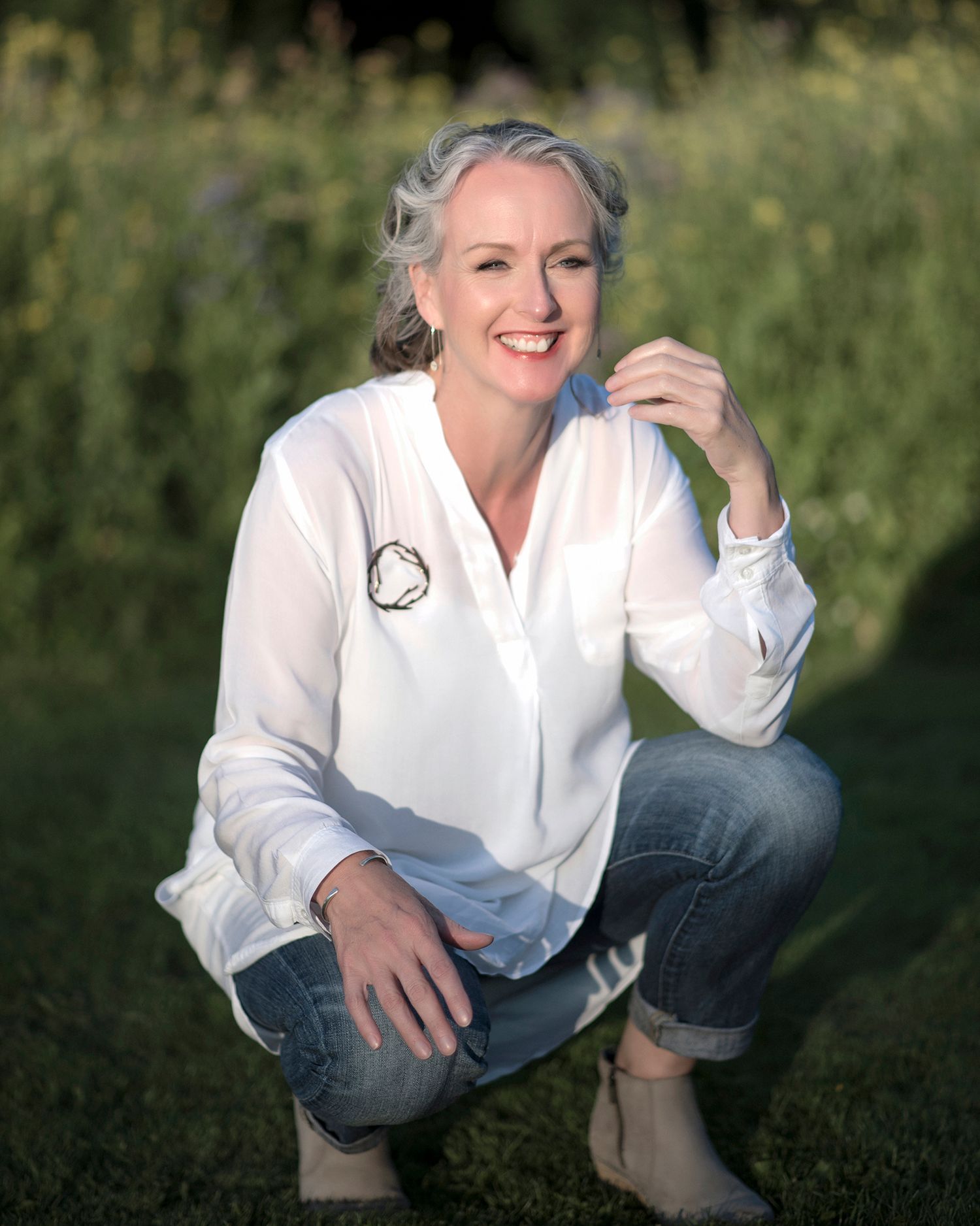 Woman in white blouse and jeans crouching outdoors in a garden, smiling warmly with gray hair and earrings.
