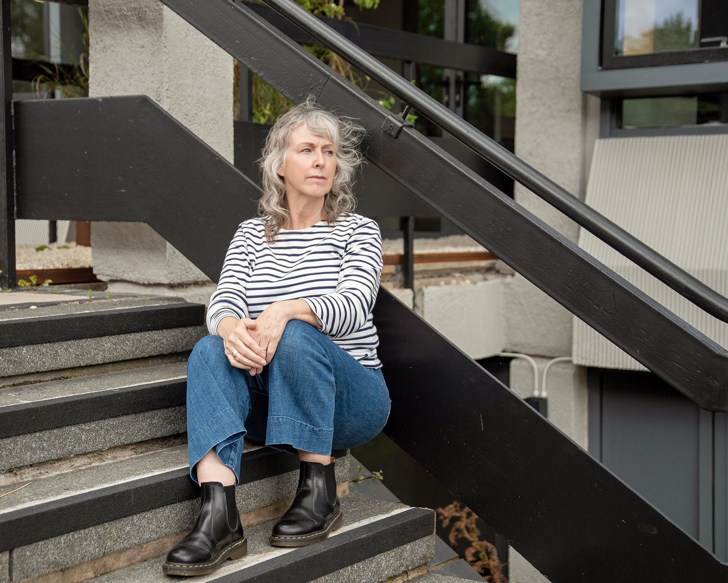 A woman with gray hair sits on outdoor steps wearing a striped shirt, jeans, and black boots.
