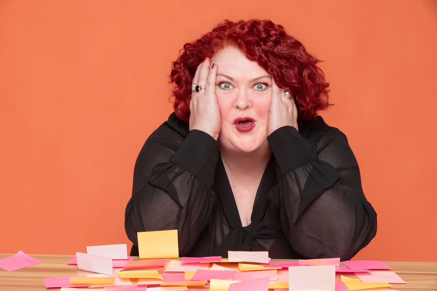 Stressed woman with curly red hair clasps her face in shock at a desk covered with scattered colorful sticky notes against an orange wall.