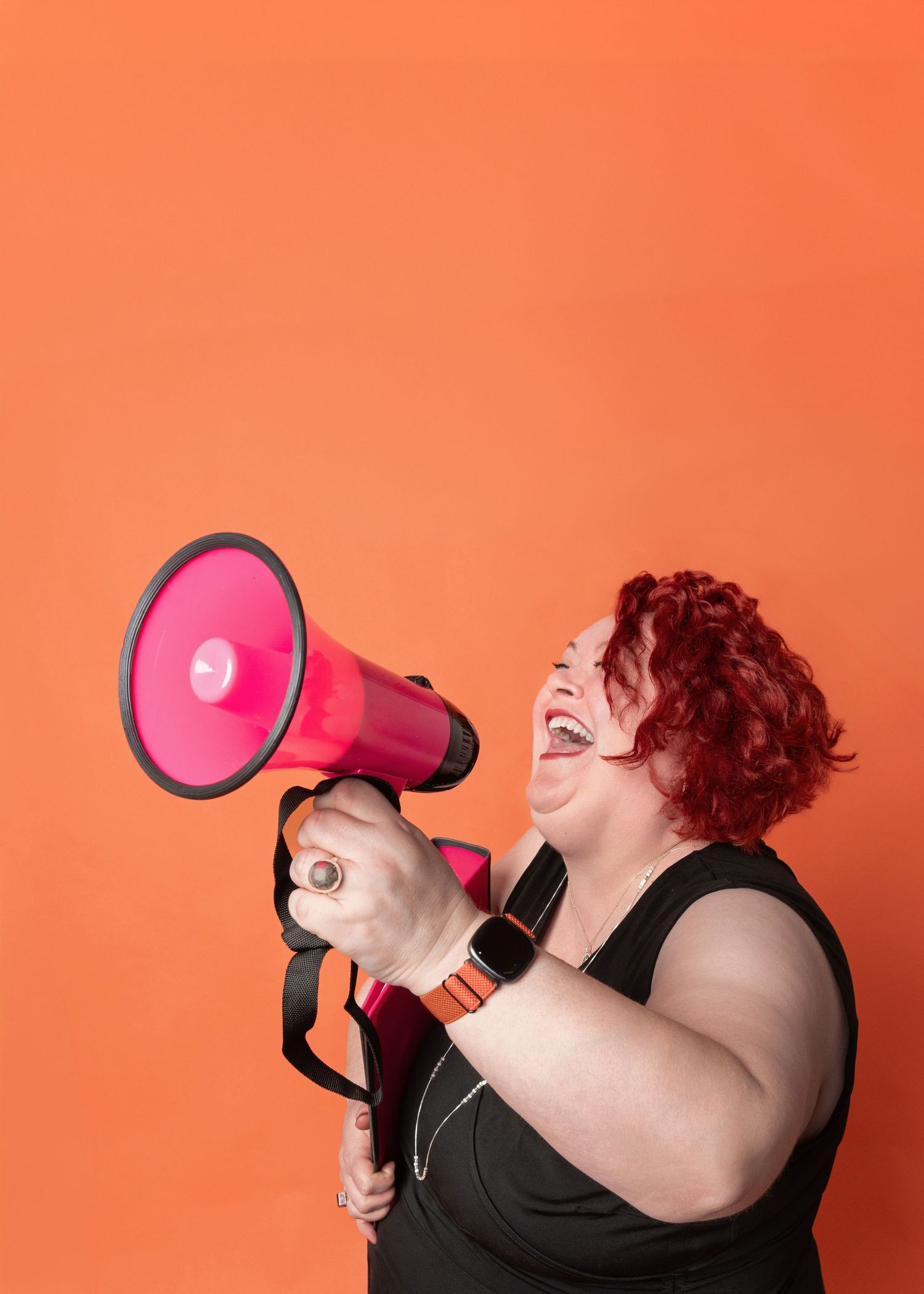 Woman with curly red hair laughs while shouting into a bright pink megaphone against a vivid orange background.
