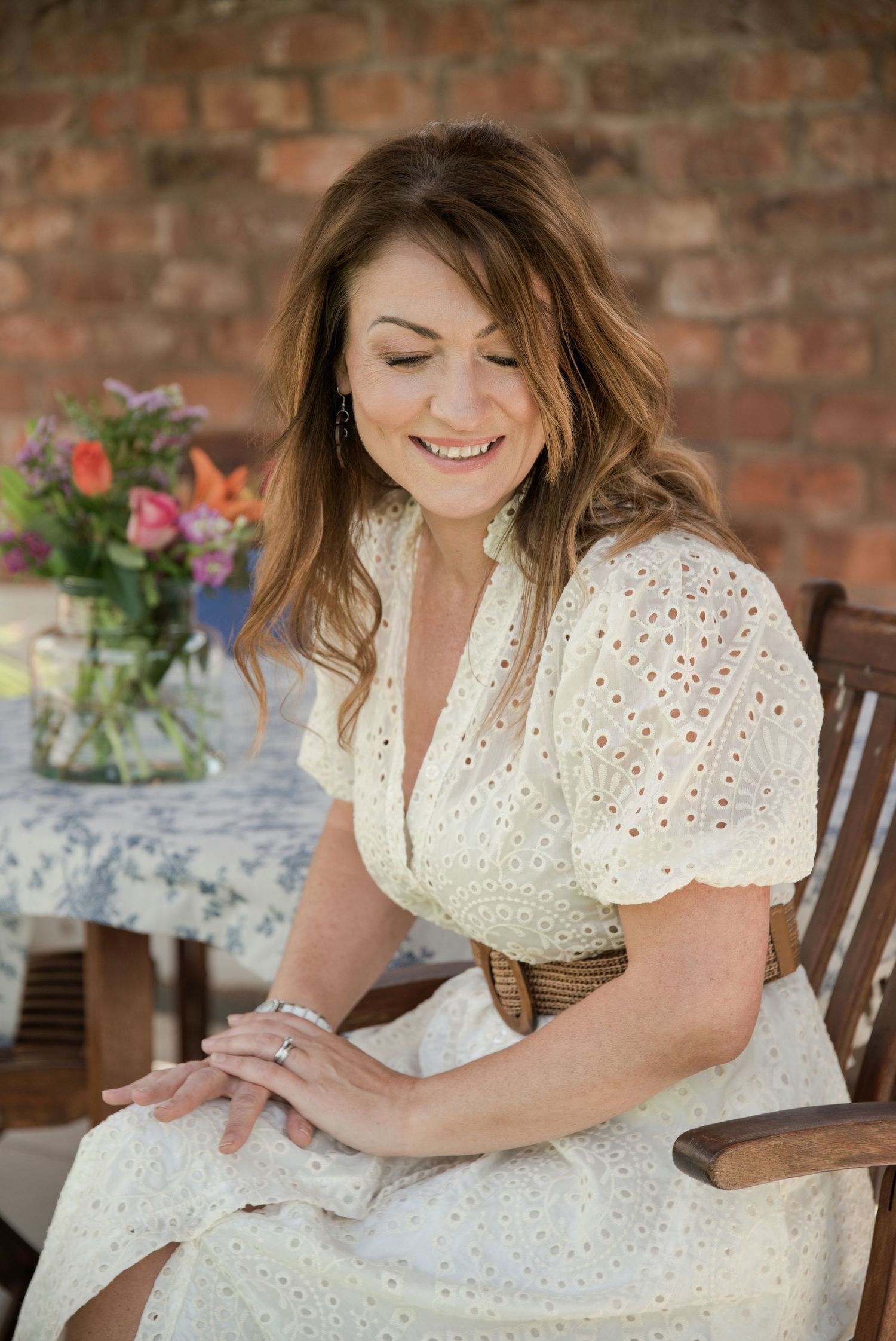 Smiling woman in white eyelet dress seated outdoors near colorful flowers and brick wall.
