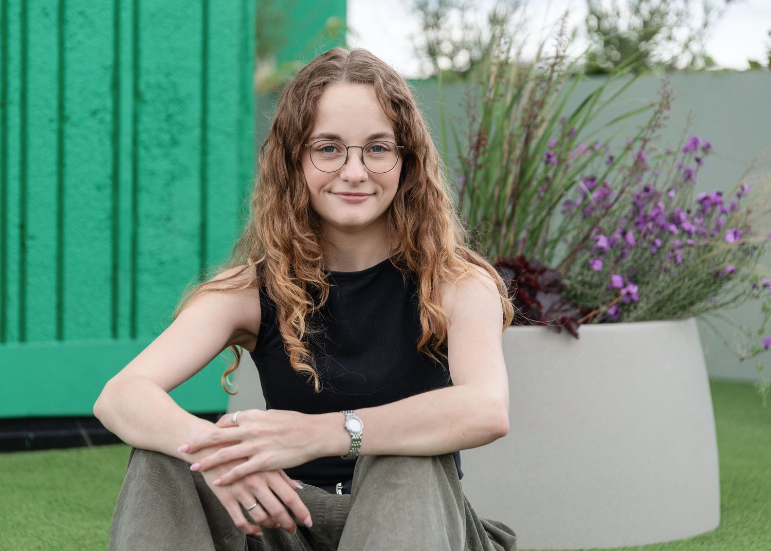 Young woman with curly hair and glasses sitting outdoors near green wall and purple flowers.