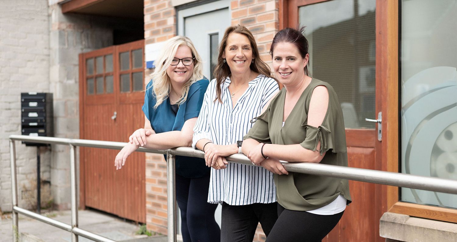 Three colleagues leaning on railing outside brick building in casual business attire.