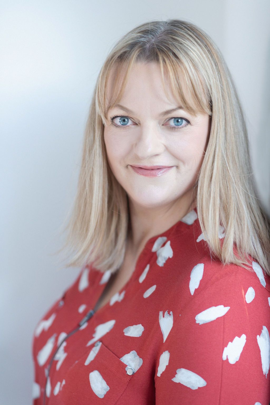 Professional headshot with blonde hair wearing a red and white patterned blouse against light background.