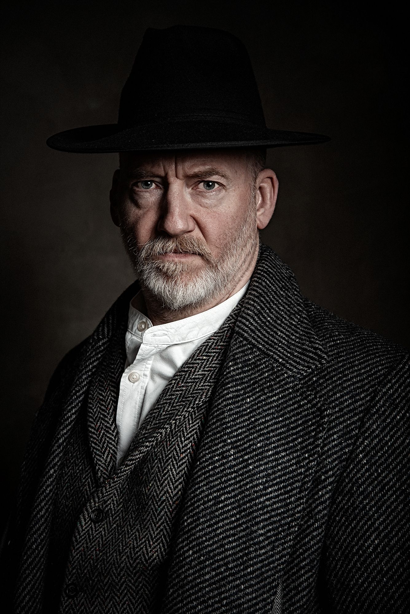 Dramatic portrait of person in wide-brimmed hat and wool coat against dark background.