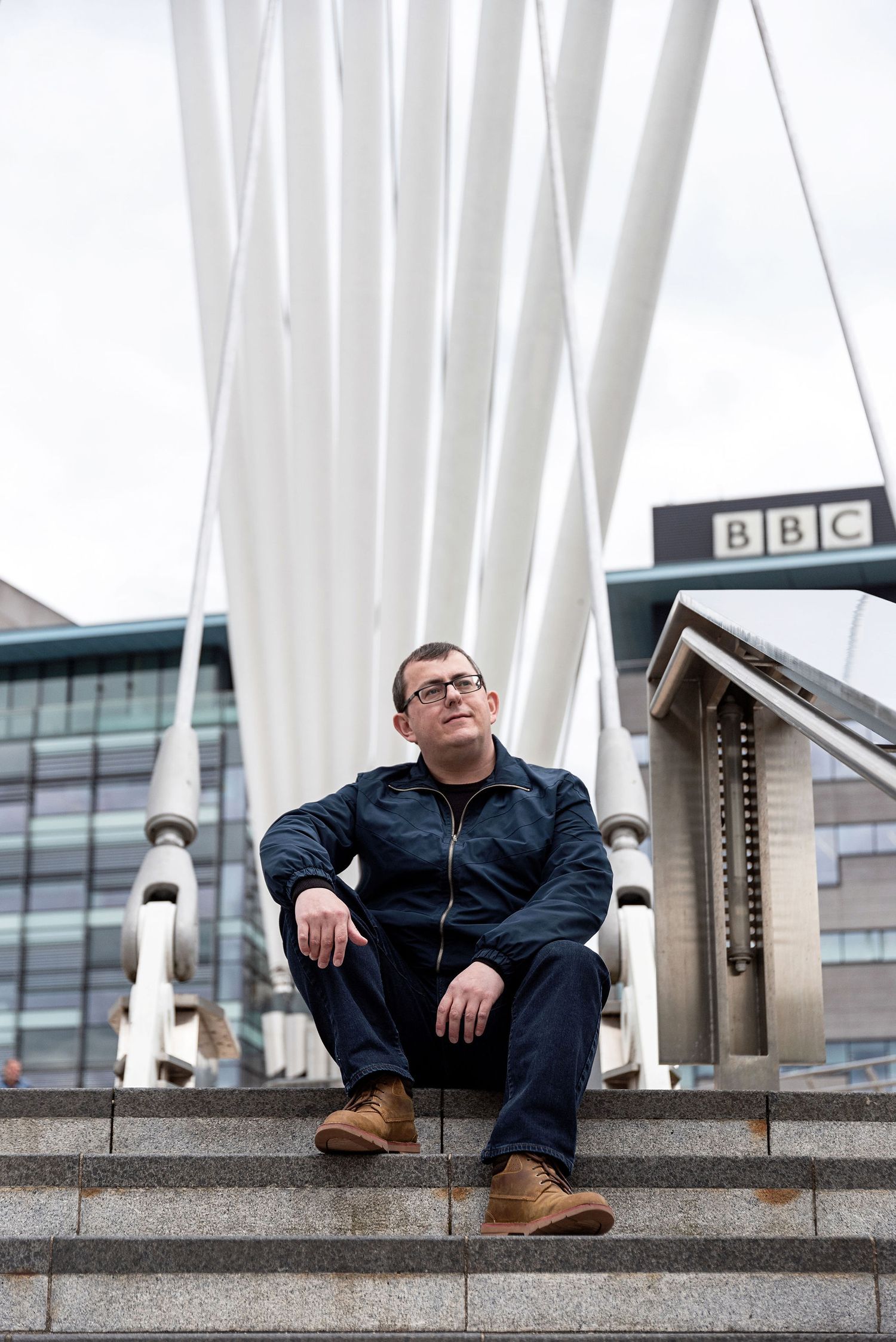 Person in navy jacket sitting on concrete steps with BBC building and white poles in background.