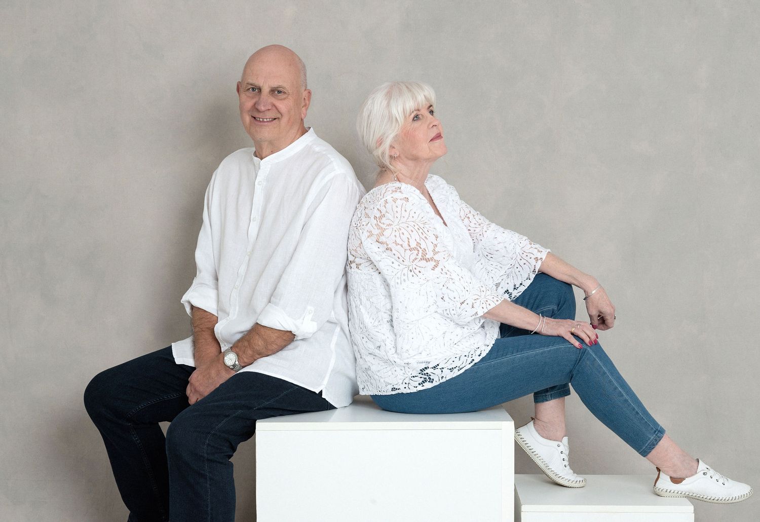 Two people in white tops and jeans sit back-to-back on white geometric blocks in a studio setting.