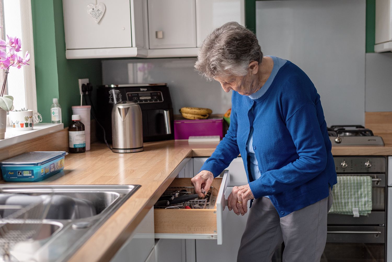 Elderly woman in blue cardigan searching through kitchen drawer in modern home kitchen.