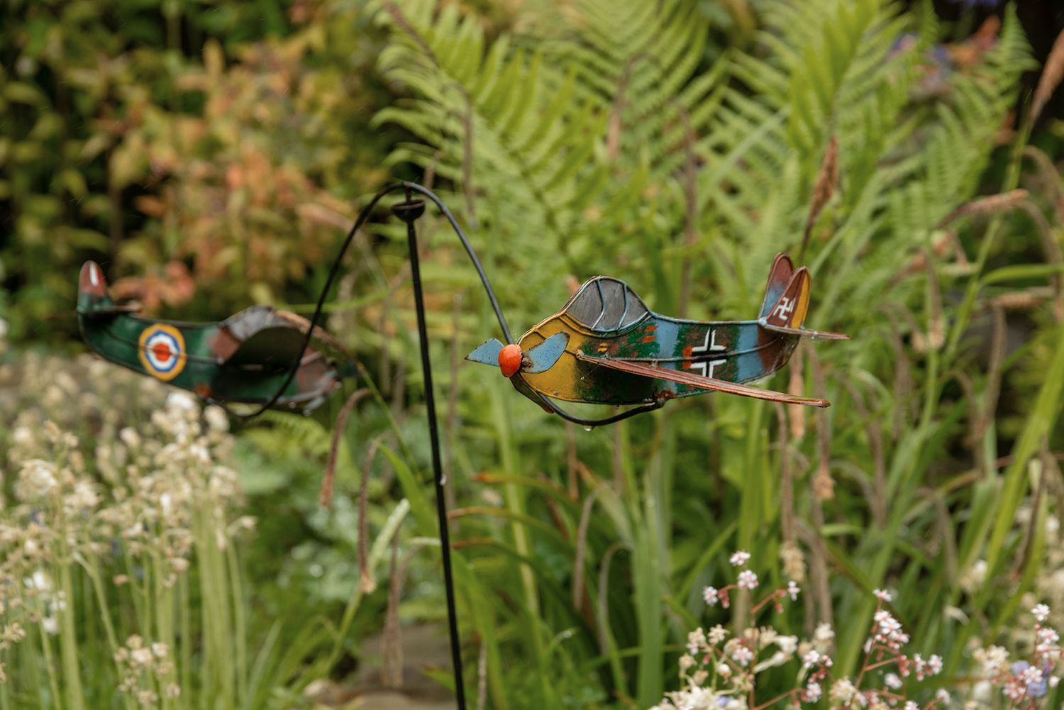 Two vintage WWII airplane garden wind spinners on metal stakes among lush green ferns and wildflowers.