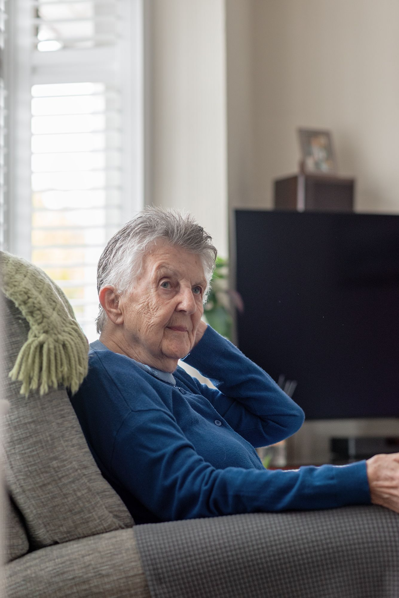 Elderly woman with grey hair sitting relaxed on a sofa in a cosy living room, wearing a blue cardigan.