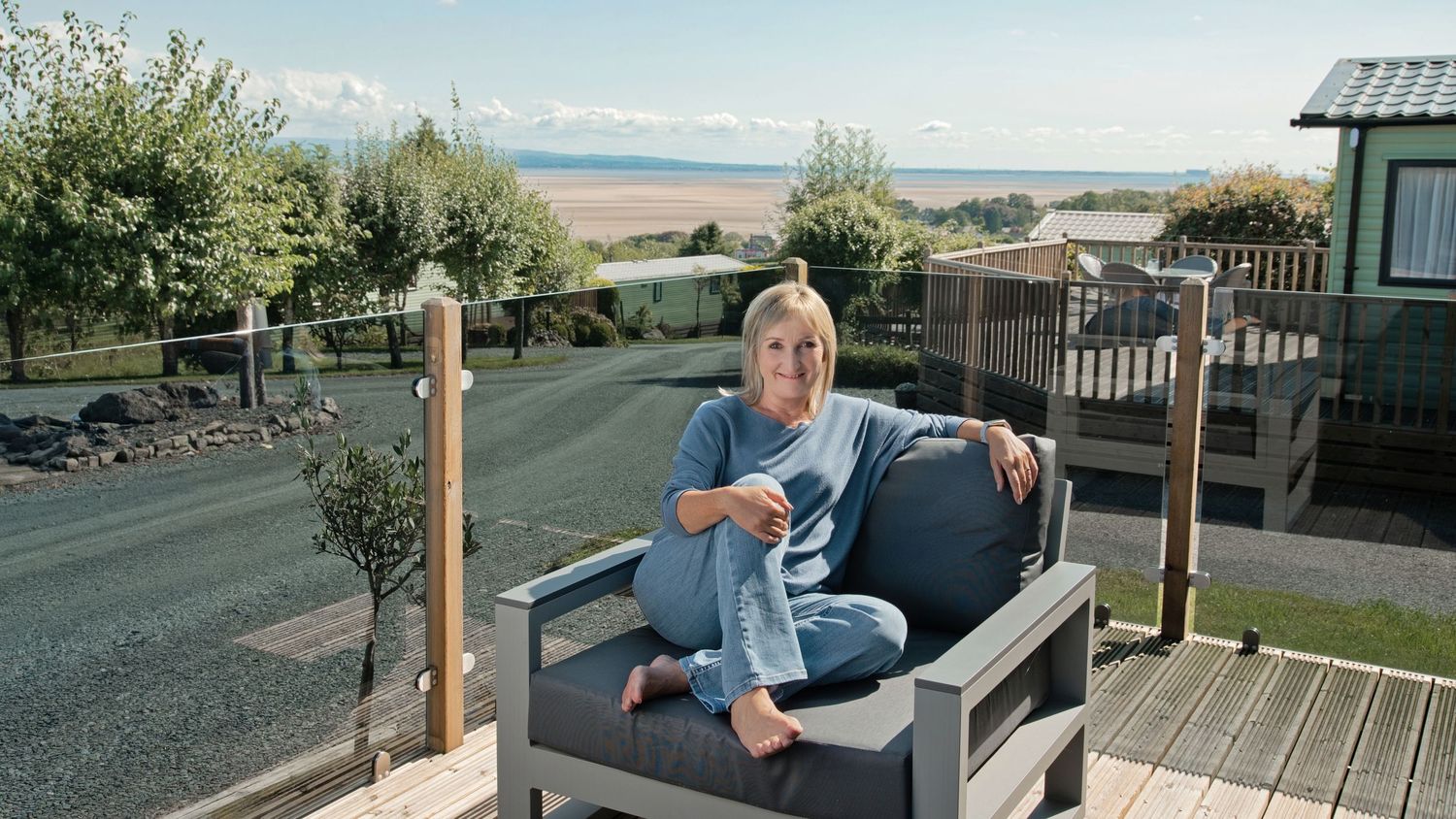 Woman relaxing on outdoor deck chair at coastal holiday park with scenic beach views.