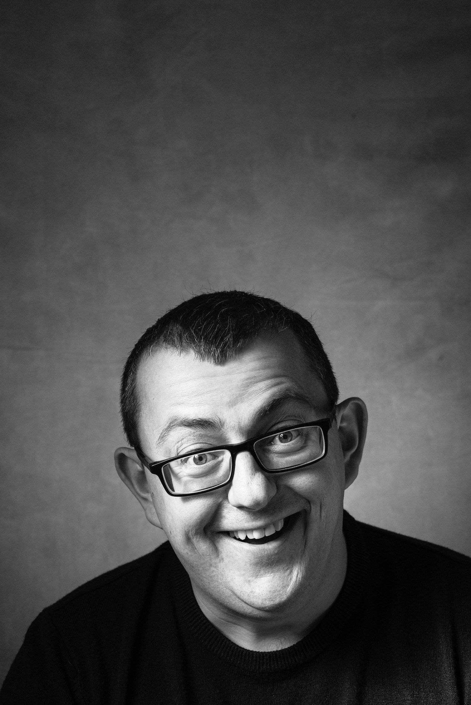 Black and white portrait of a smiling man wearing rectangular glasses, making a funny face against a gray background.