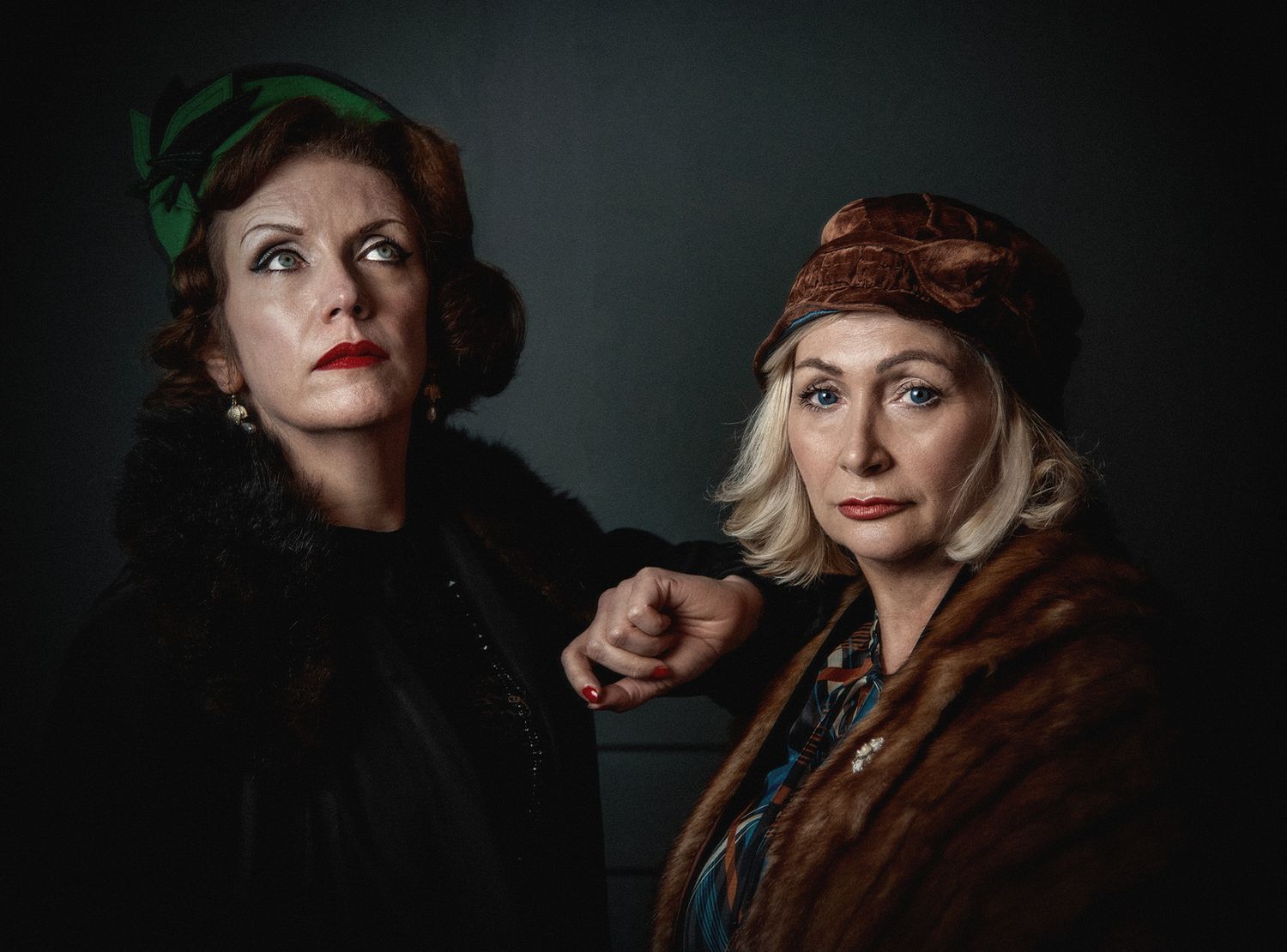 Two women in vintage-style clothing and headpieces pose dramatically against dark background.