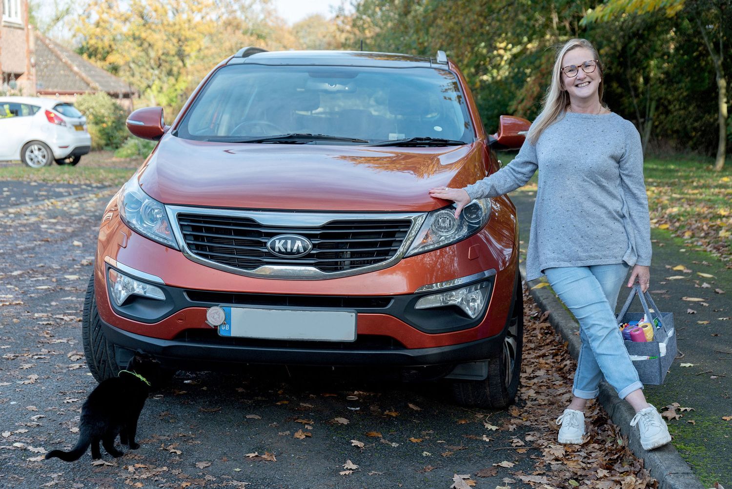 Person standing next to red Kia SUV with black dog in autumn setting.