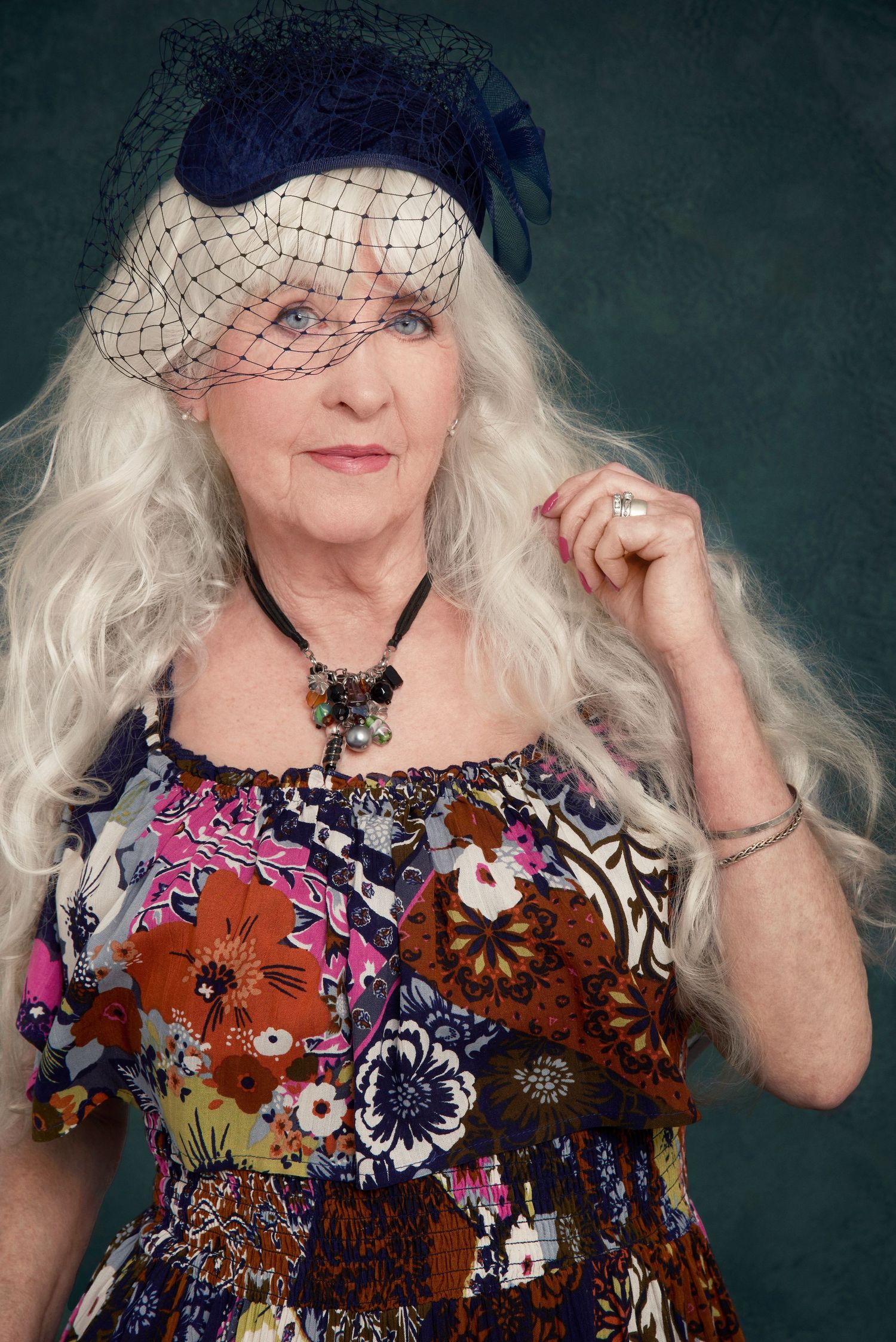 Person wearing a floral dress and navy fascinator with veil poses elegantly against a dark background.