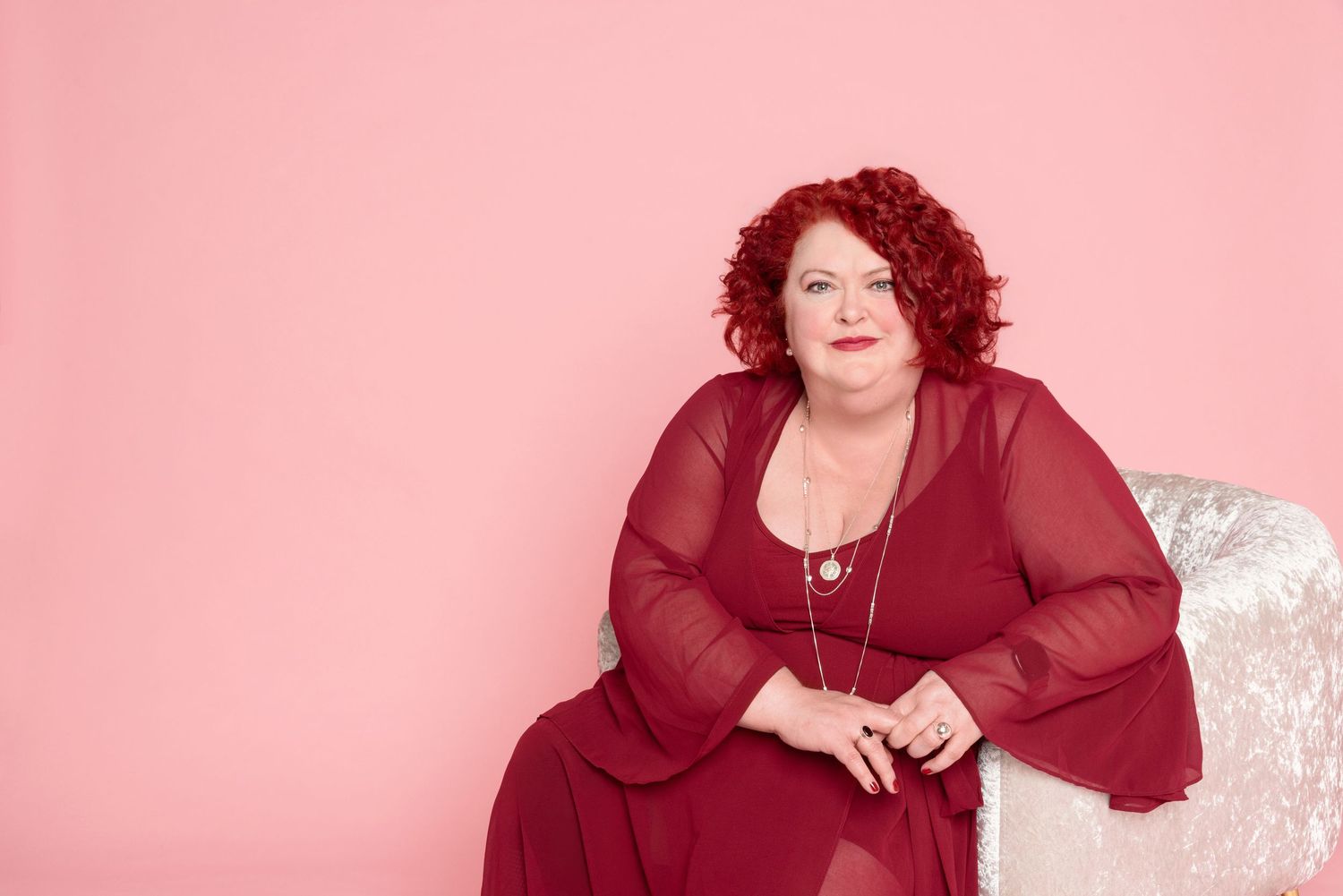 Woman with curly red hair in a flowing burgundy sheer-sleeved dress sits on a sparkly white chair against a pink background.
