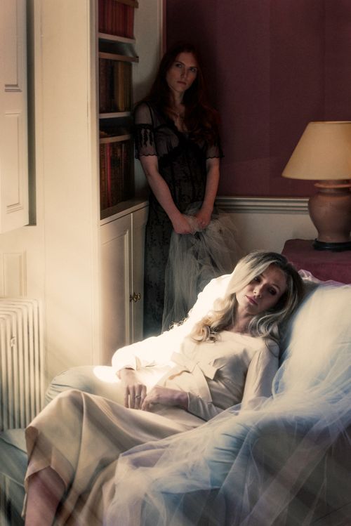Two women in a dimly lit bedroom, one reclining on a bed in white, another standing by a bookcase.