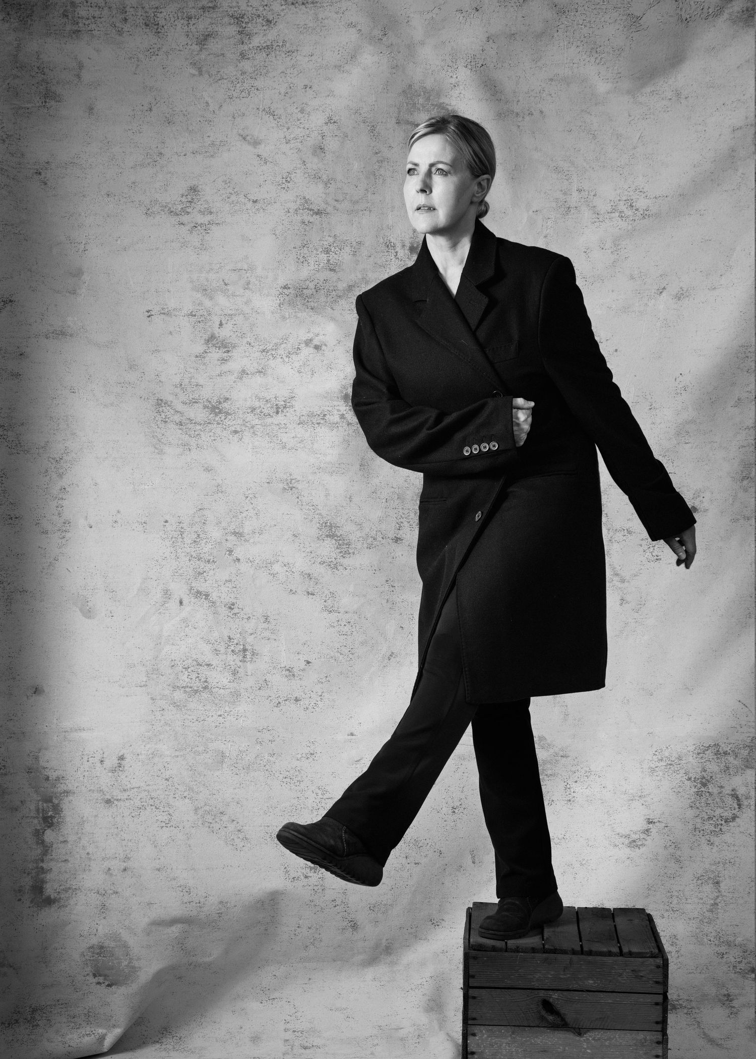 Black and white portrait of a person in a dark oversized coat standing on a wooden box against a textured background.