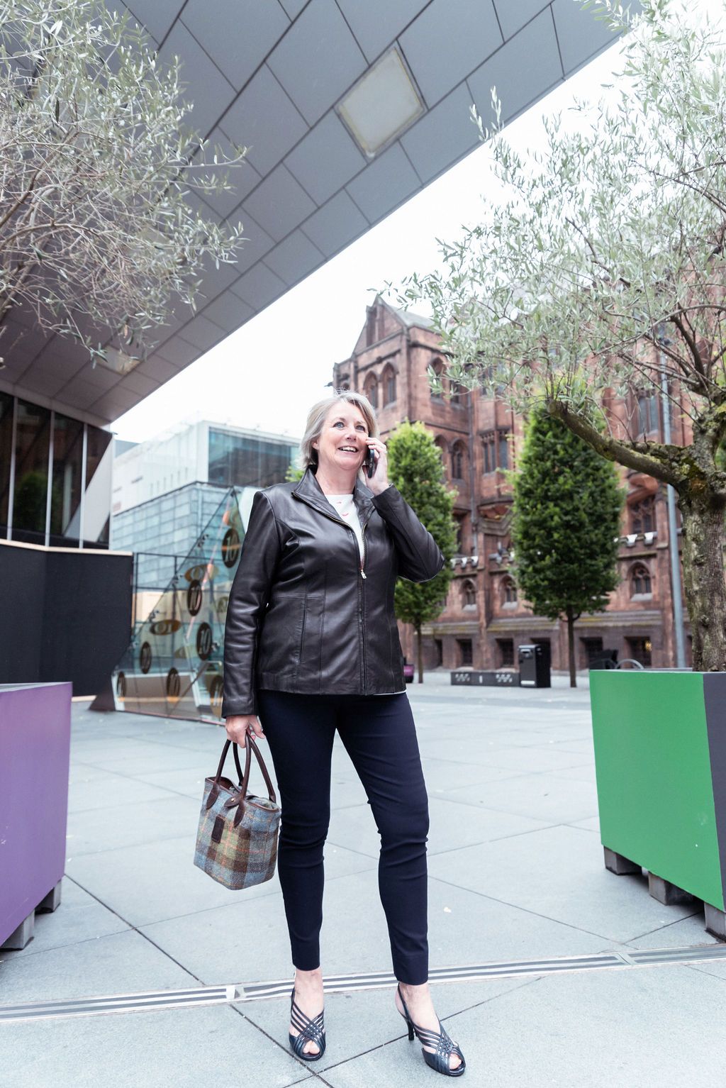 Professional woman in leather jacket talking on phone while carrying handbag outdoors near modern building.
