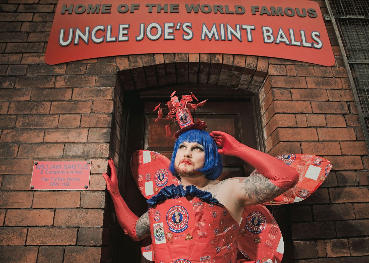 Person in red costume with blue hair poses outside Uncle Joe's Mint Balls factory in Wigan, England.