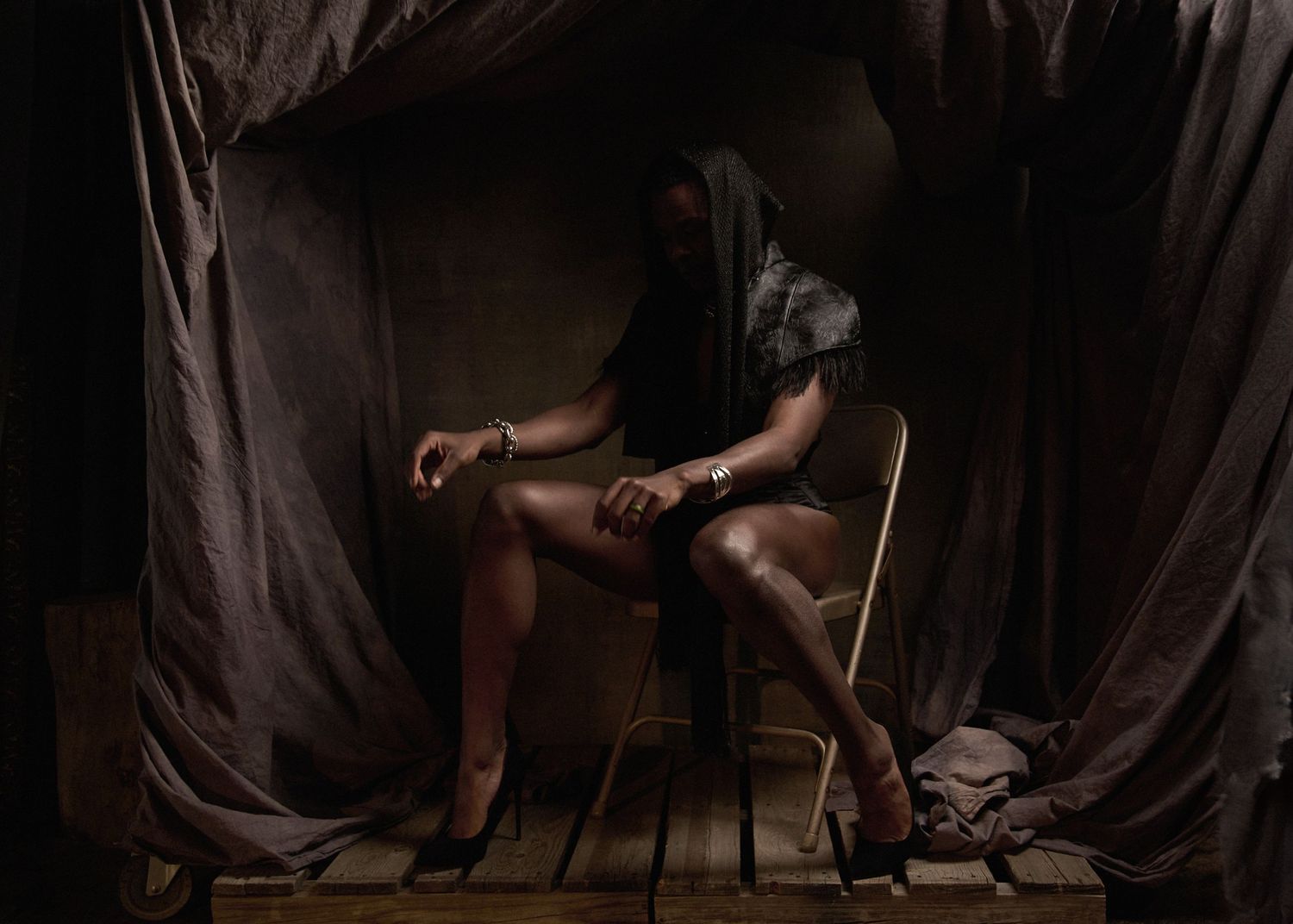 Dramatic low-key photograph of figure seated in chair against dark fabric backdrop.
