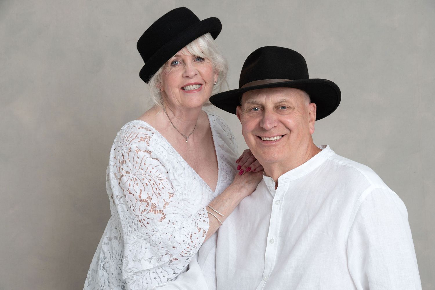Couple wearing matching black hats and white outfits pose together against a gray background.