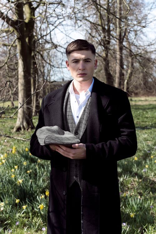 Young man in dark overcoat and plaid vest holding a flat cap, standing in a park with daffodils and bare trees.