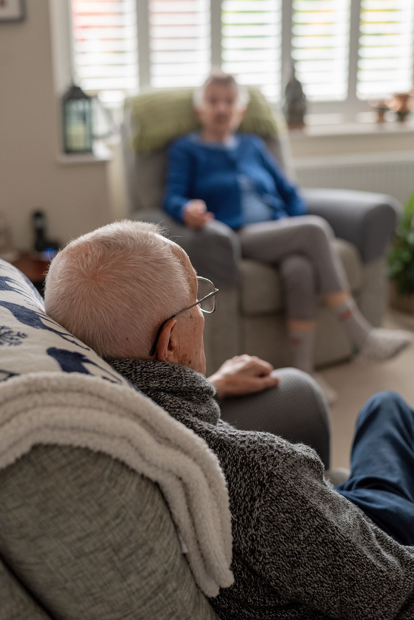 Elderly couple relaxing in armchairs in a cozy living room, senior man with glasses in foreground, woman in blue top behind.