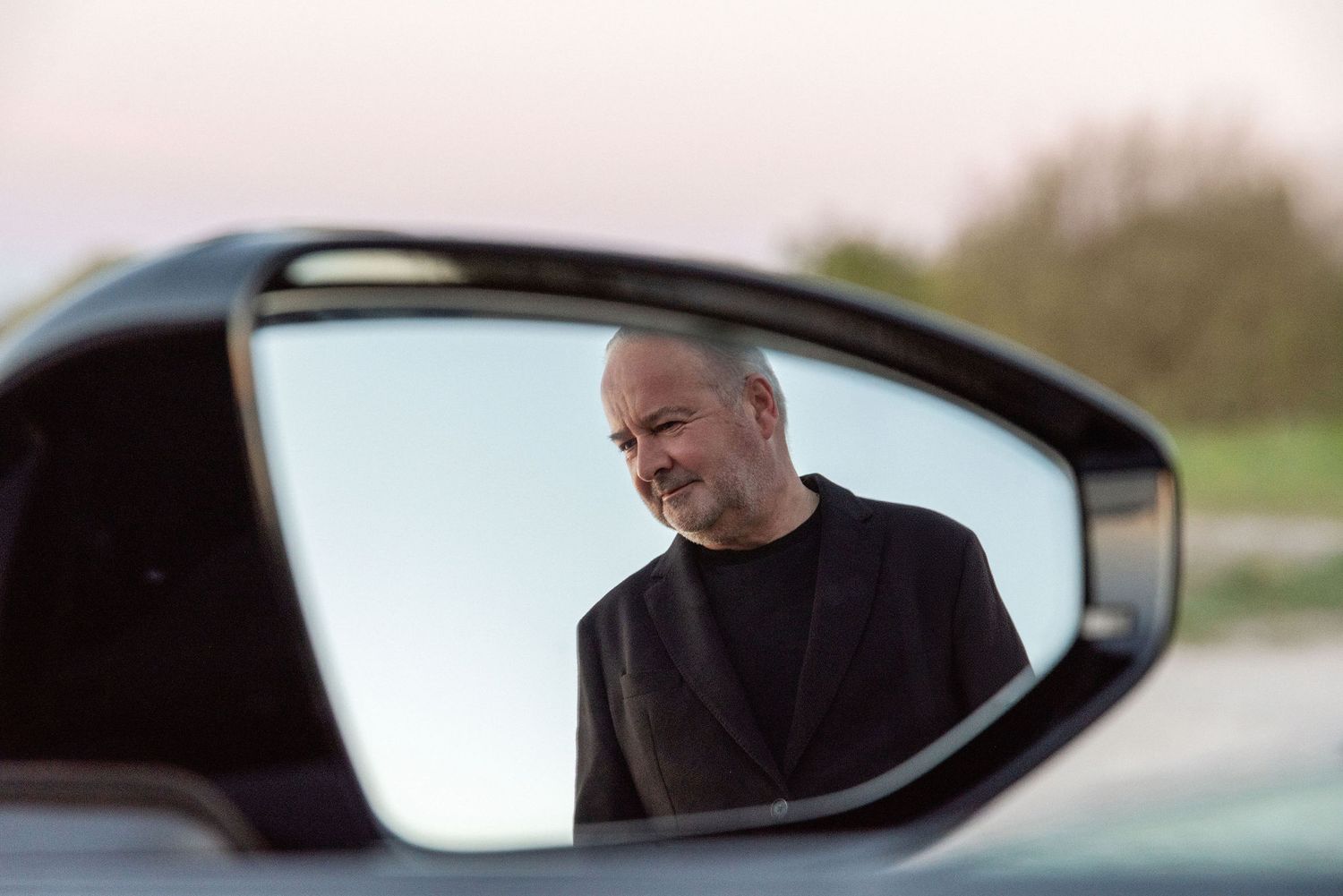 Man reflected in car side mirror smiling outdoors at dusk.