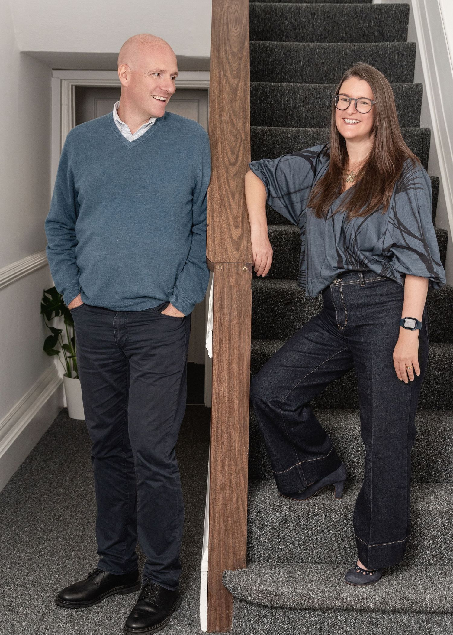 A man and woman in coordinating blue outfits pose on a staircase, smiling in a modern home interior.