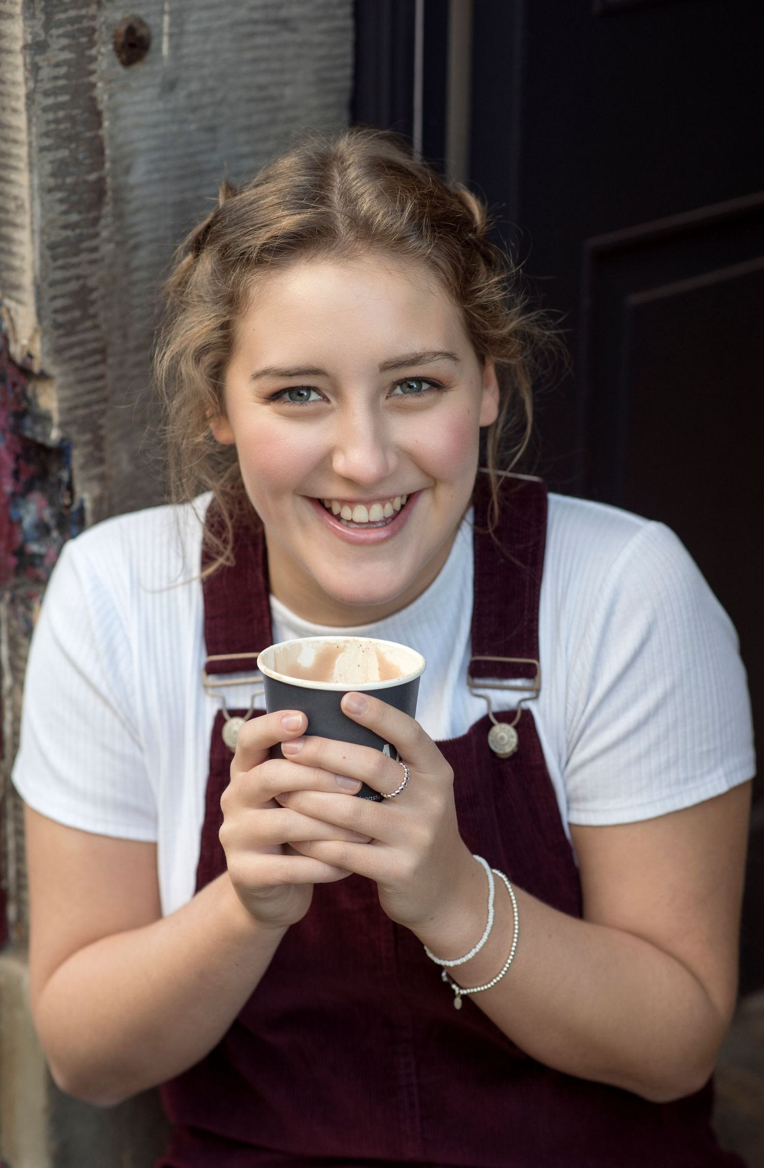 Smiling young woman holding a takeaway coffee cup wearing burgundy dungarees and a white top outdoors.