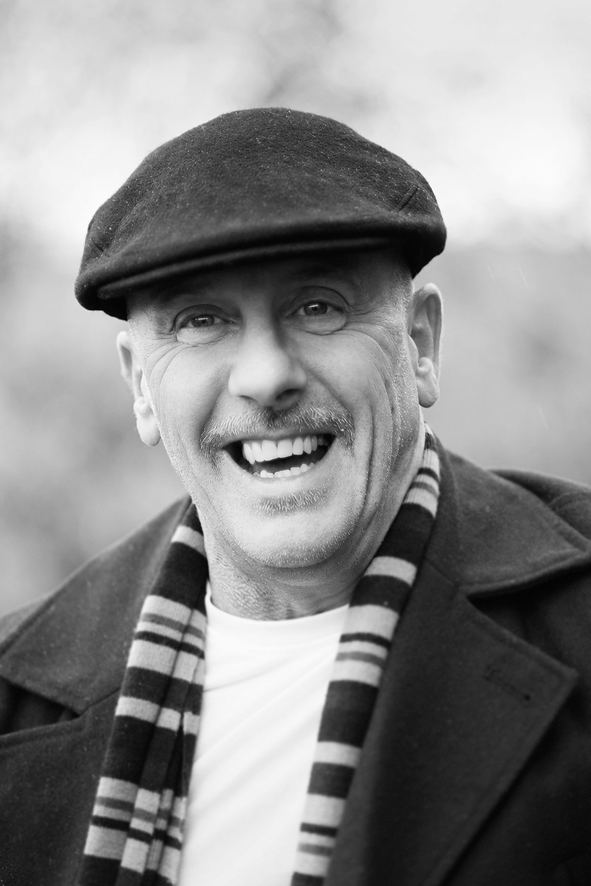 Smiling older man with mustache wearing flat cap and striped scarf in black and white portrait photo.