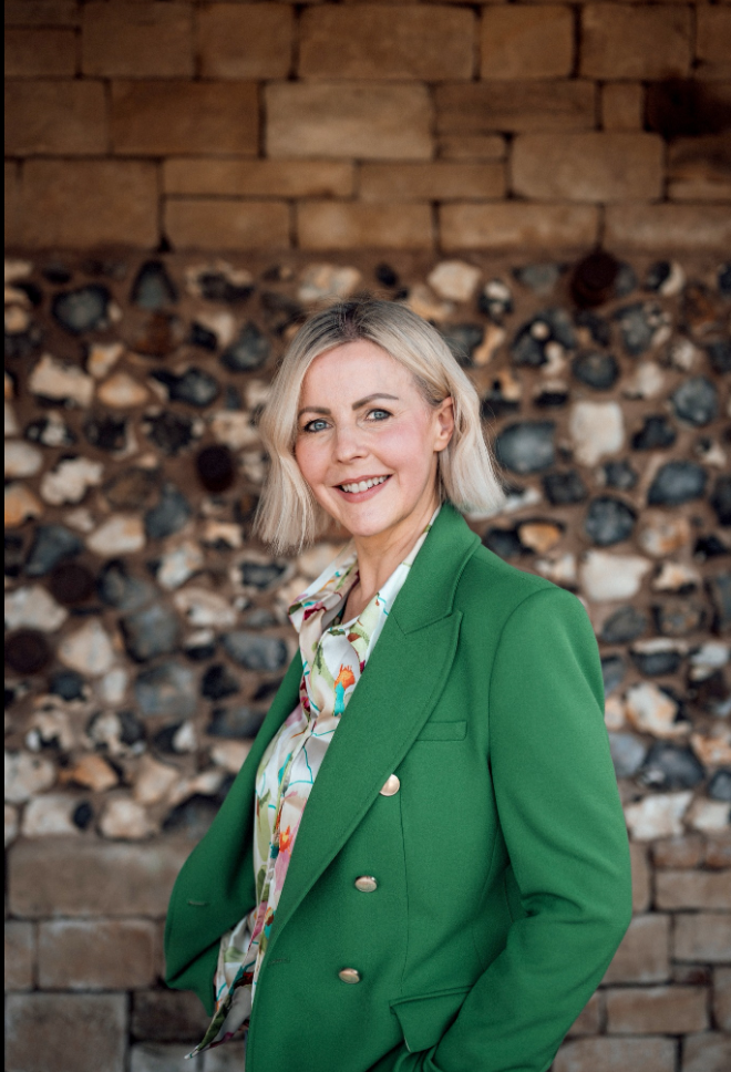 Woman in green blazer and floral blouse smiling confidently in front of a rustic stone wall backdrop.