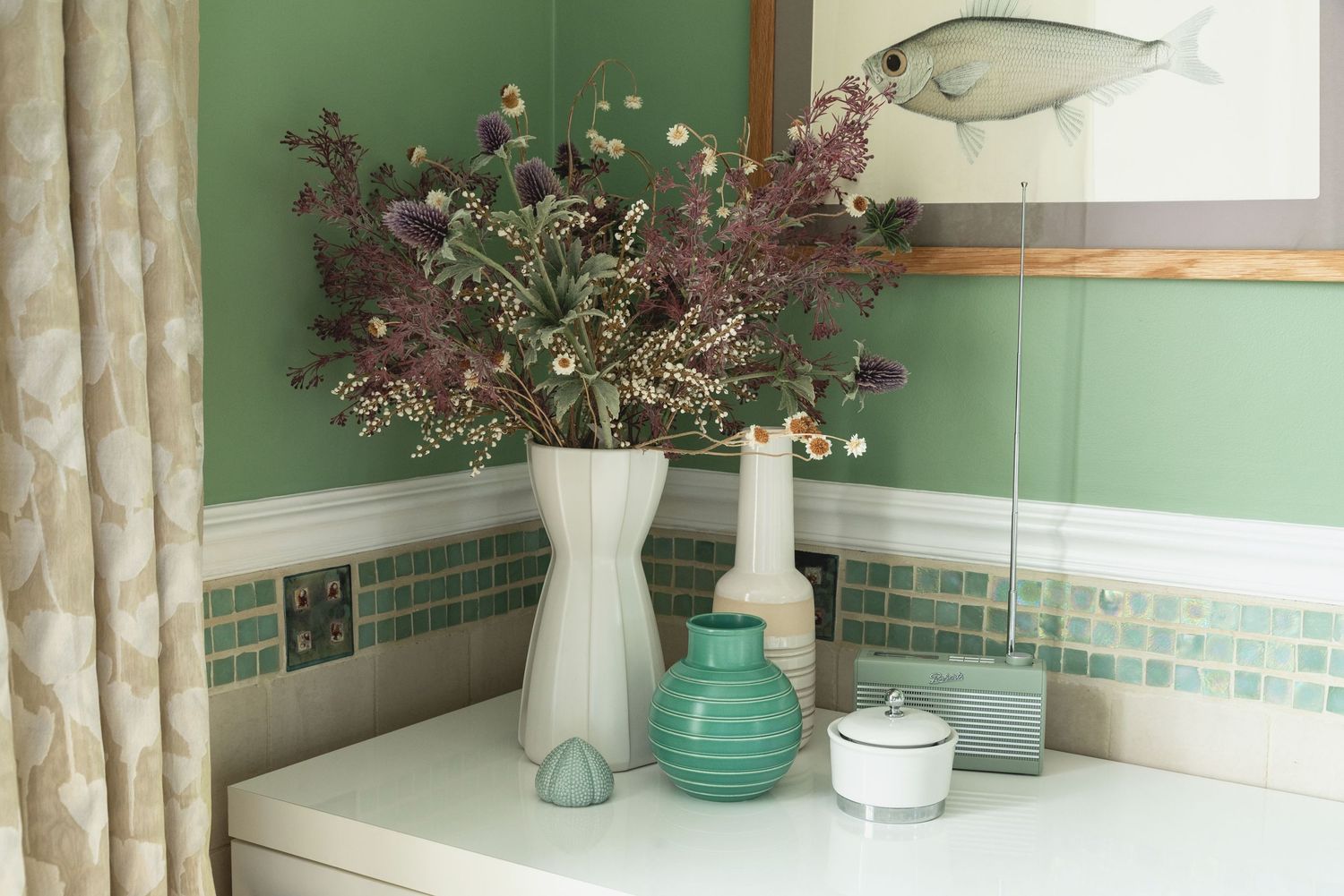 Green bathroom shelf with white vase of dried wildflowers, ceramic vases, and a fish artwork on sage green wall.
