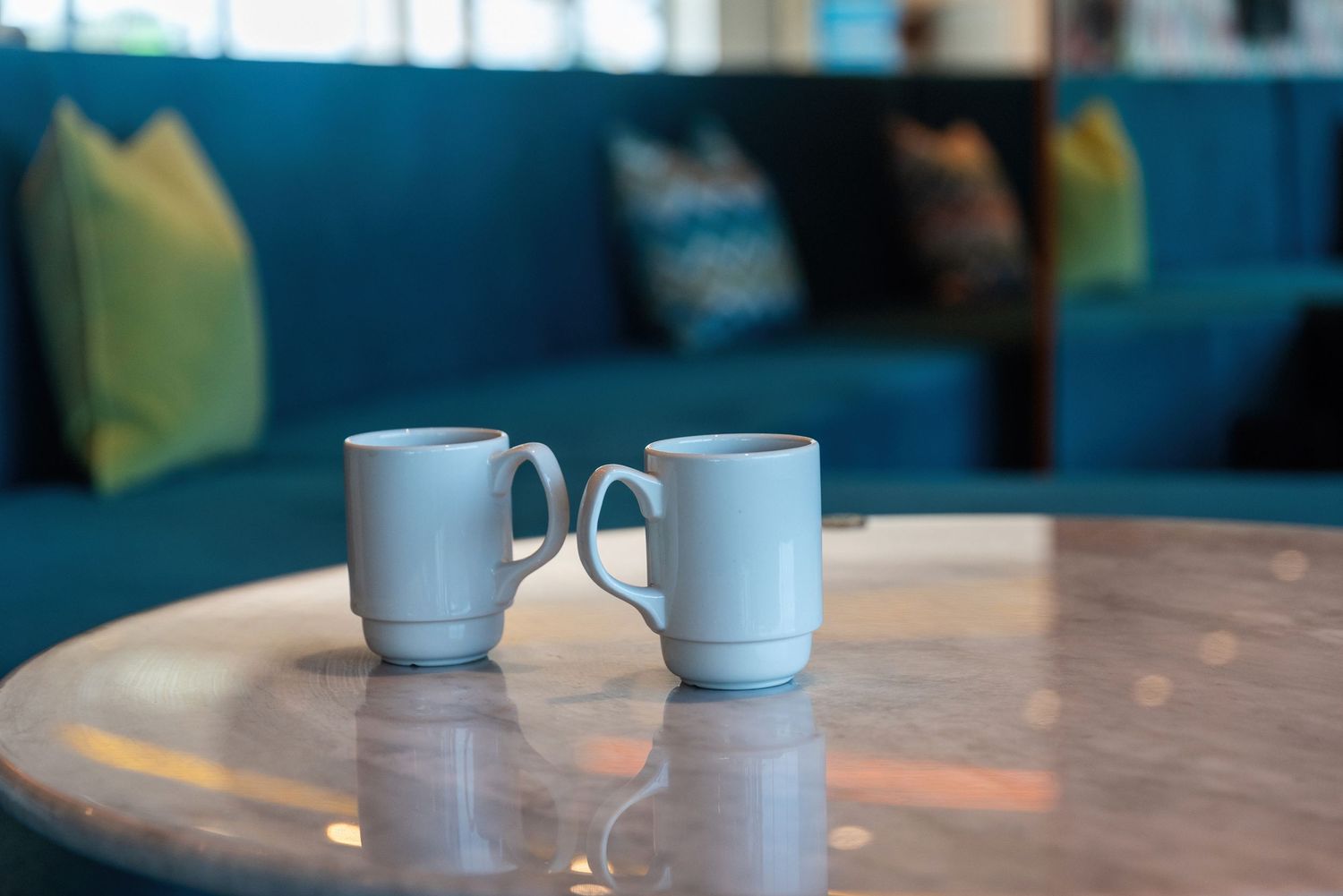 Two white ceramic coffee mugs on round table with blue seating in background.