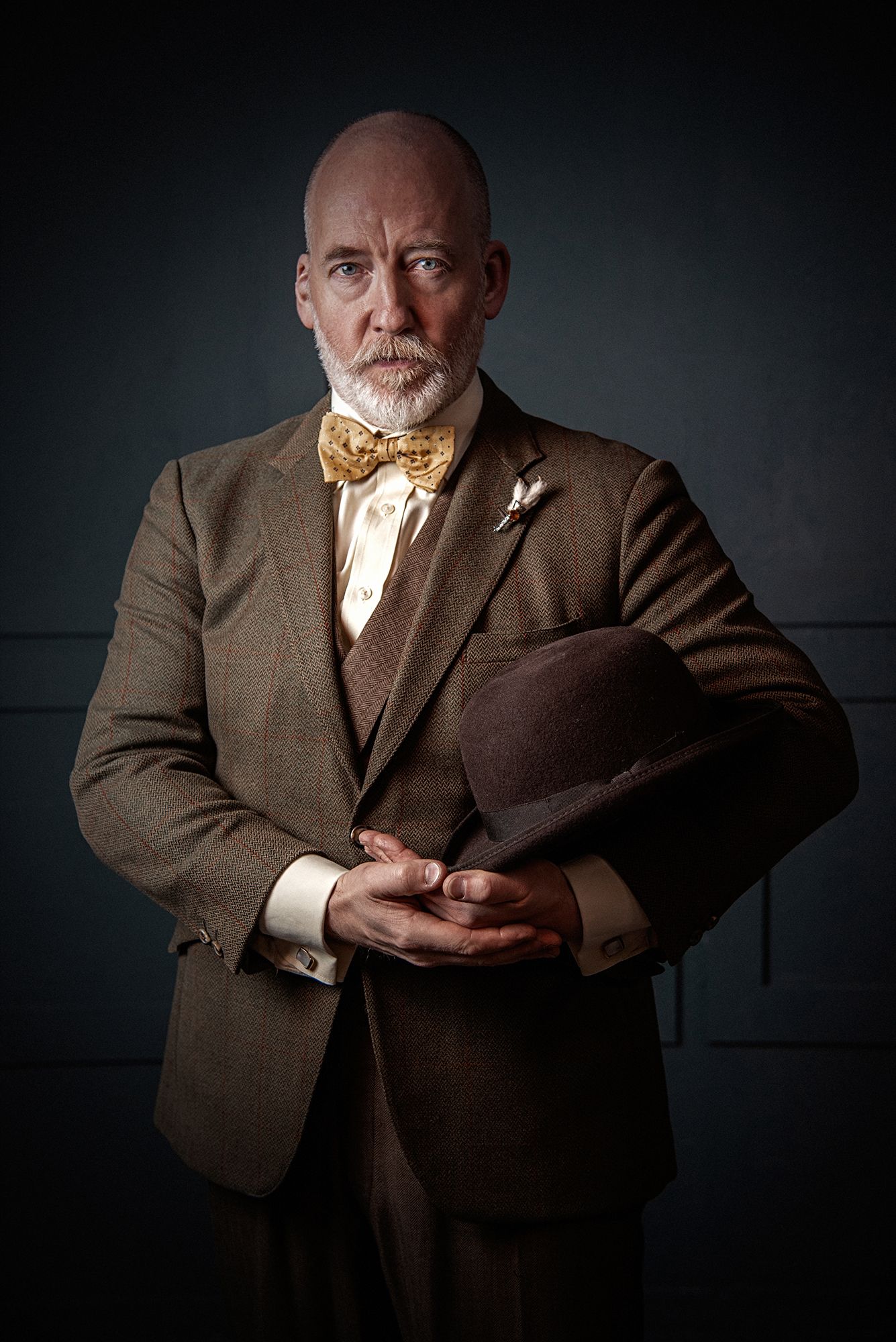 Vintage-style portrait of person in brown suit and bowtie holding hat against dark background.