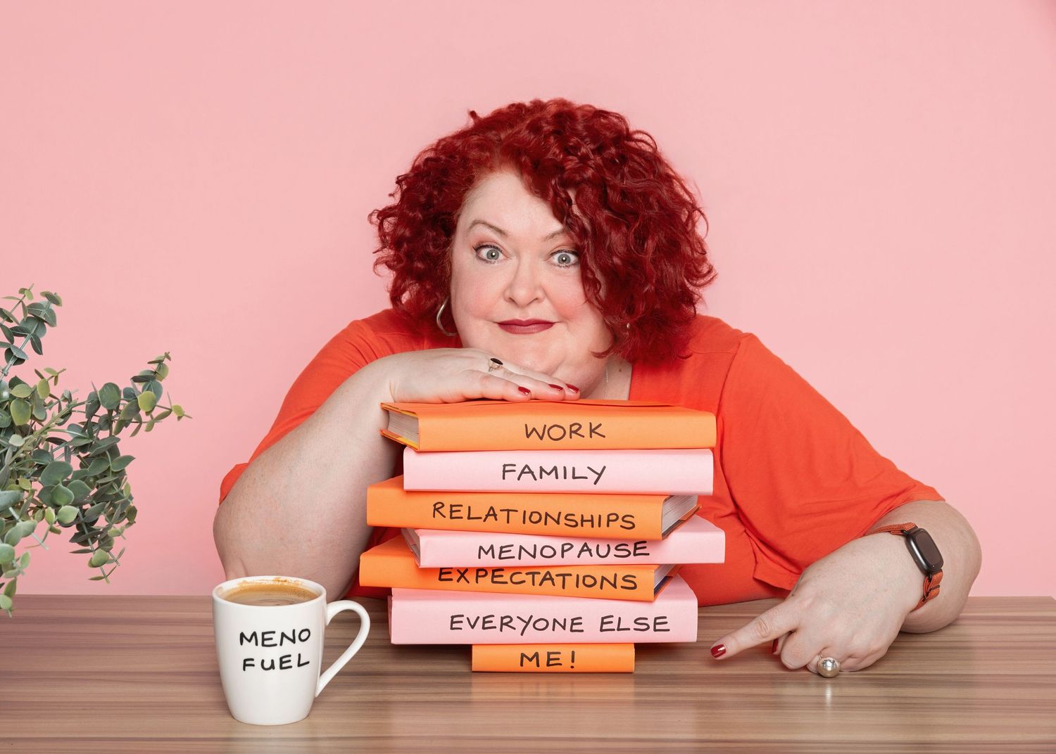 Smiling woman with curly red hair rests her chin on a stack of books labeled work, family, menopause and me beside a Meno Fuel mug.